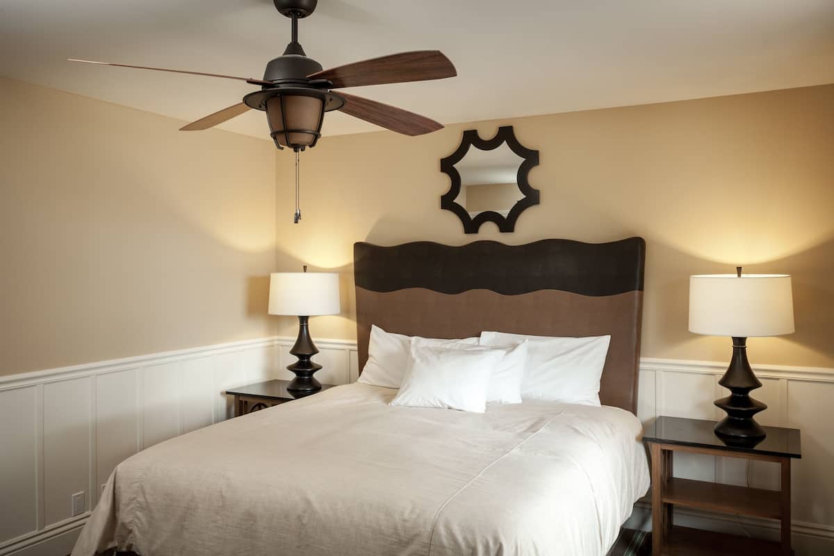 A neatly made bed with a wooden headboard, two bedside lamps, a ceiling fan, and a decorative mirror on the wall.