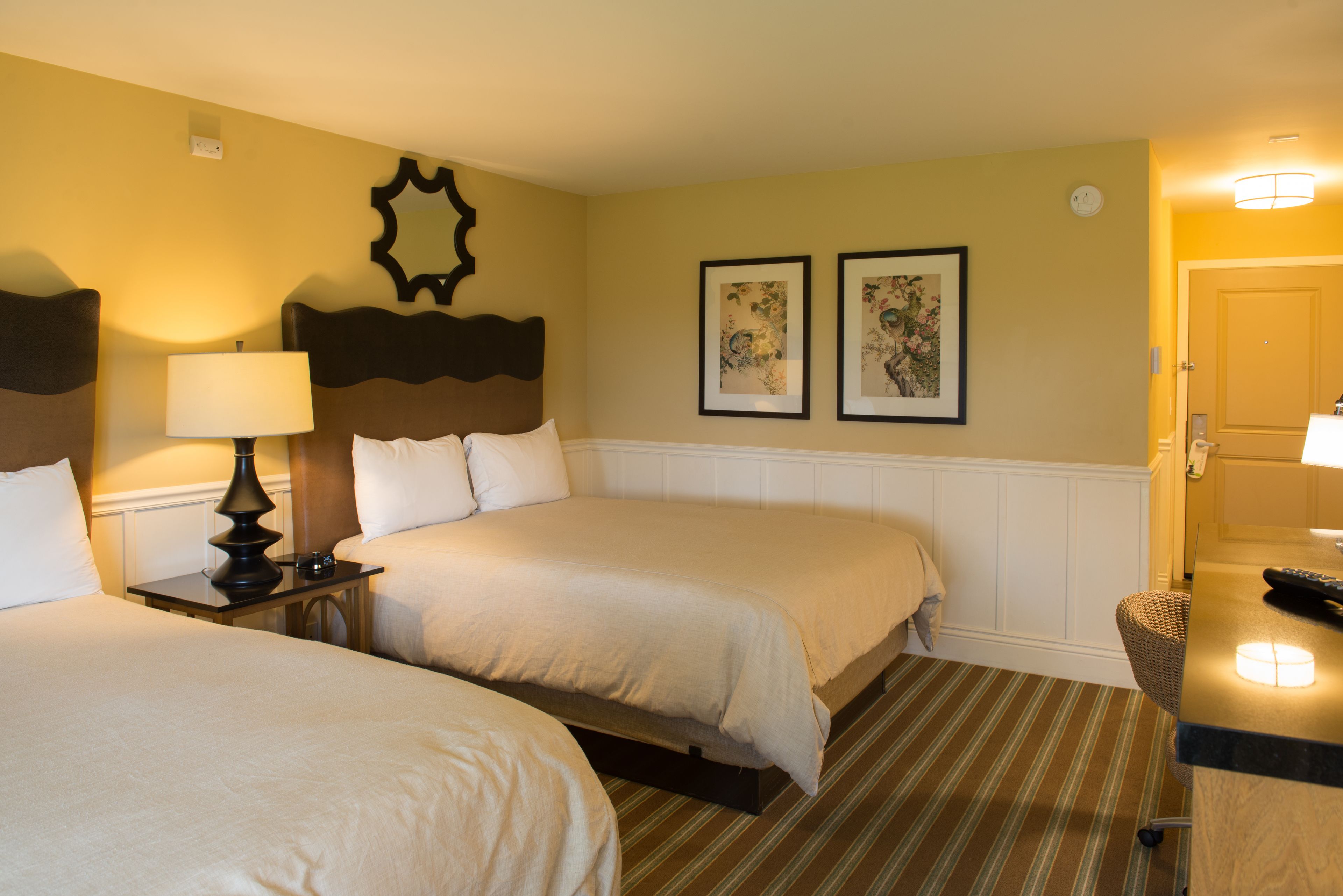 A hotel room with two beds, a nightstand with a lamp, a framed mirror, and two framed pictures on the wall.