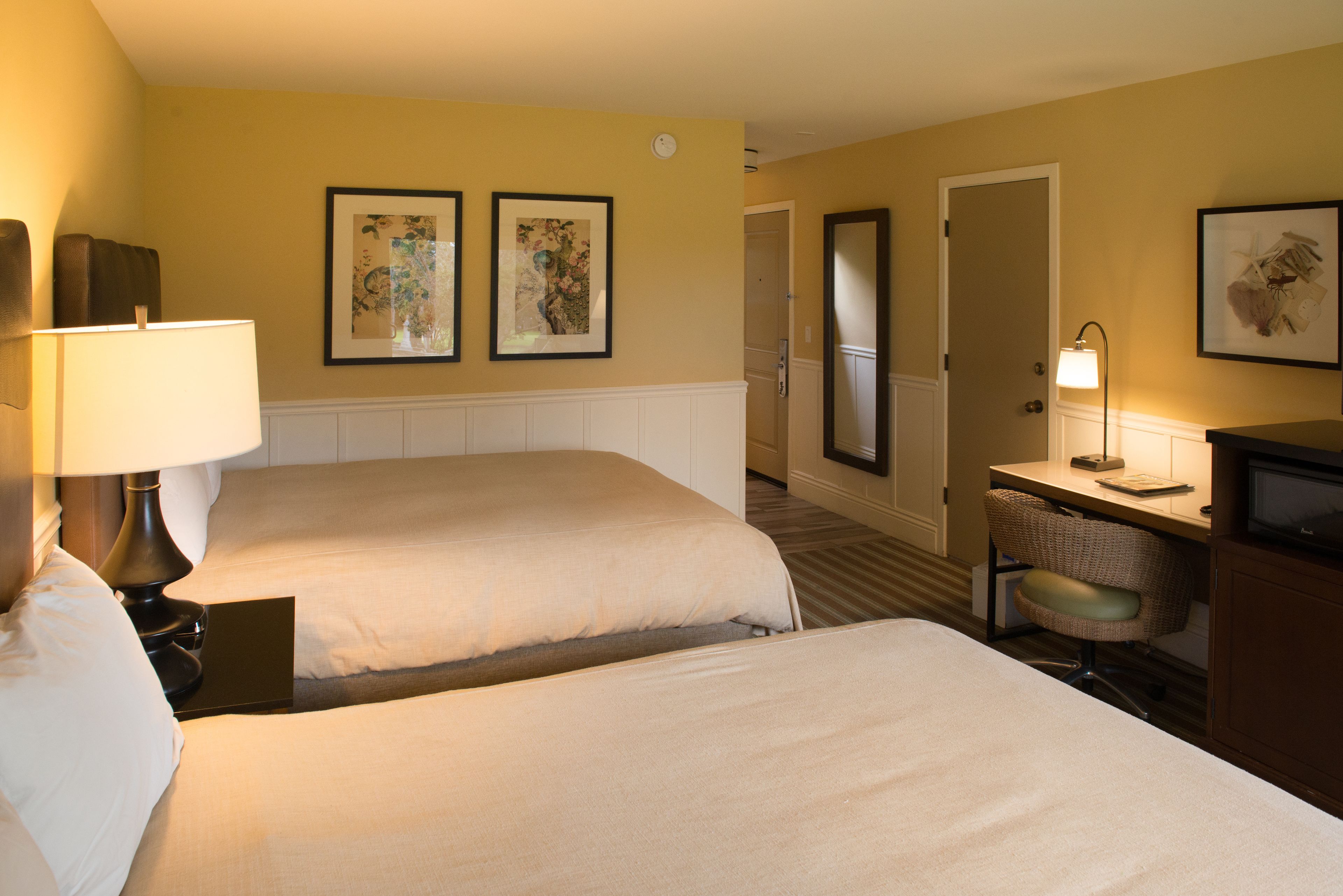 A hotel room with two beds, a desk, a chair, and framed artwork on the walls.