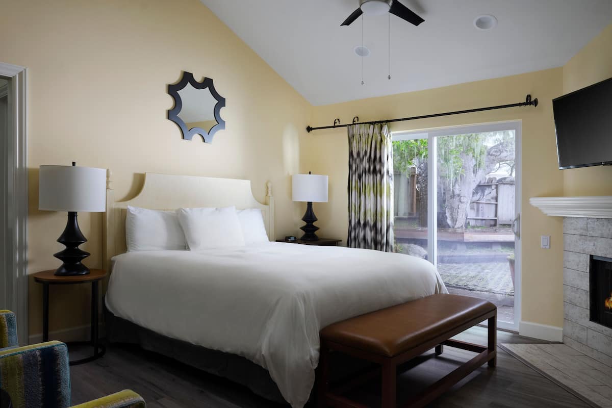 A bedroom with a bed, two bedside tables with lamps, a bench, a fireplace, and a TV mounted on the wall.