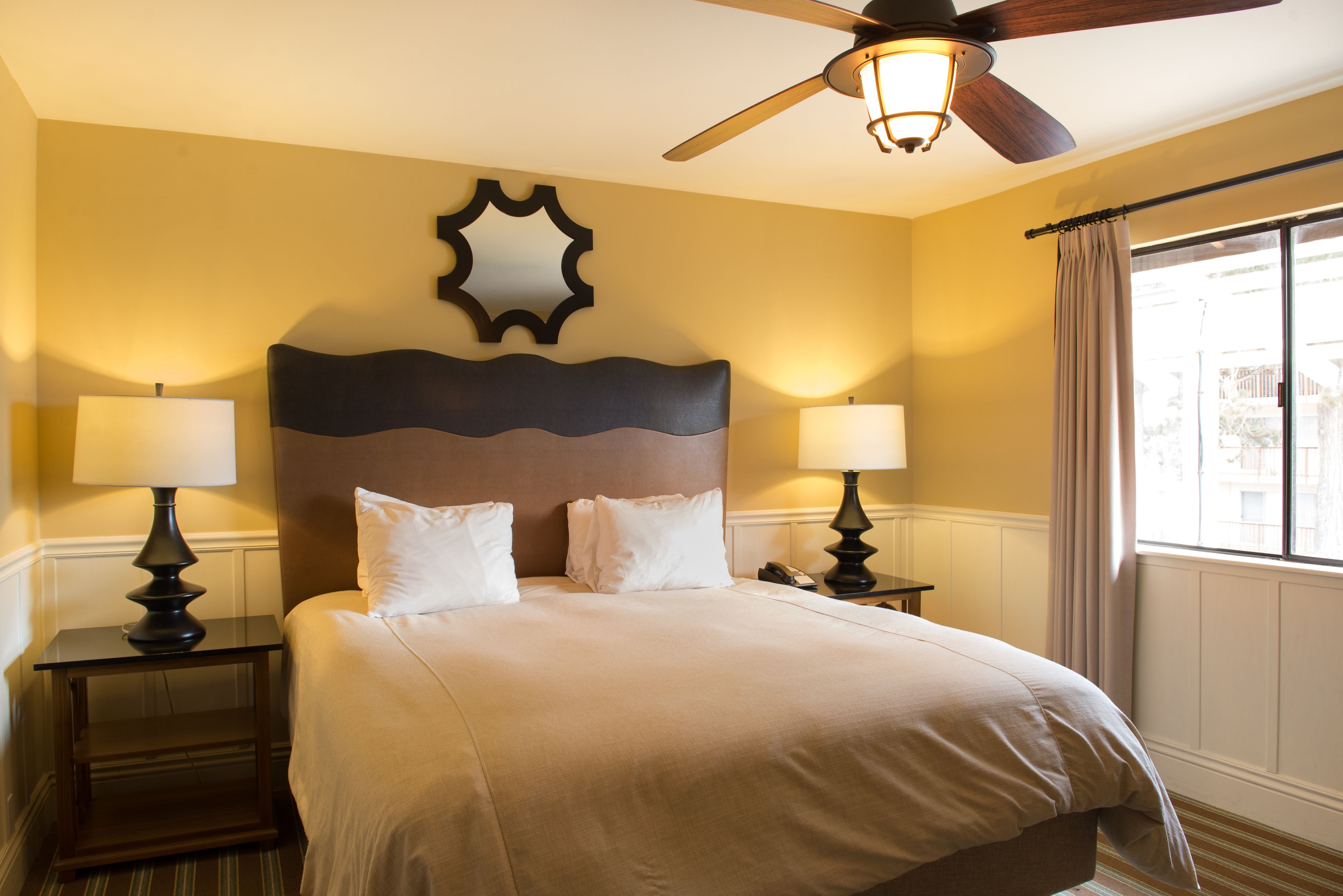 Une chambre d’hôtel avec un grand lit, deux tables de chevet avec des lampes, un ventilateur de plafond, un miroir décoratif au mur et une fenêtre avec des rideaux.