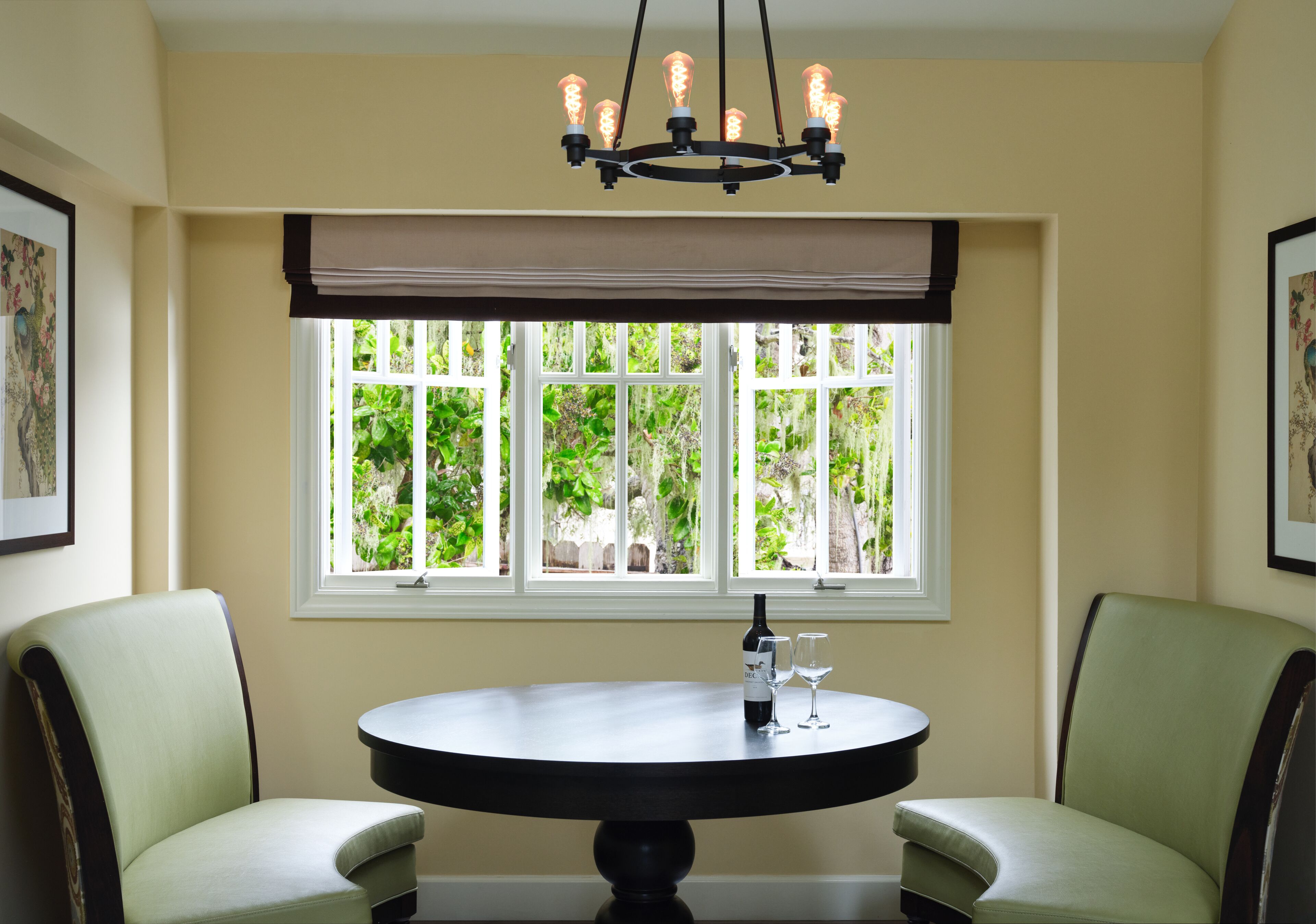 Un comedor con una mesa redonda, dos sillas, una lámpara de araña y una ventana con vistas a la vegetación.