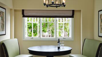 A dining area with a round table, two chairs, a chandelier, and a window with a view of greenery.