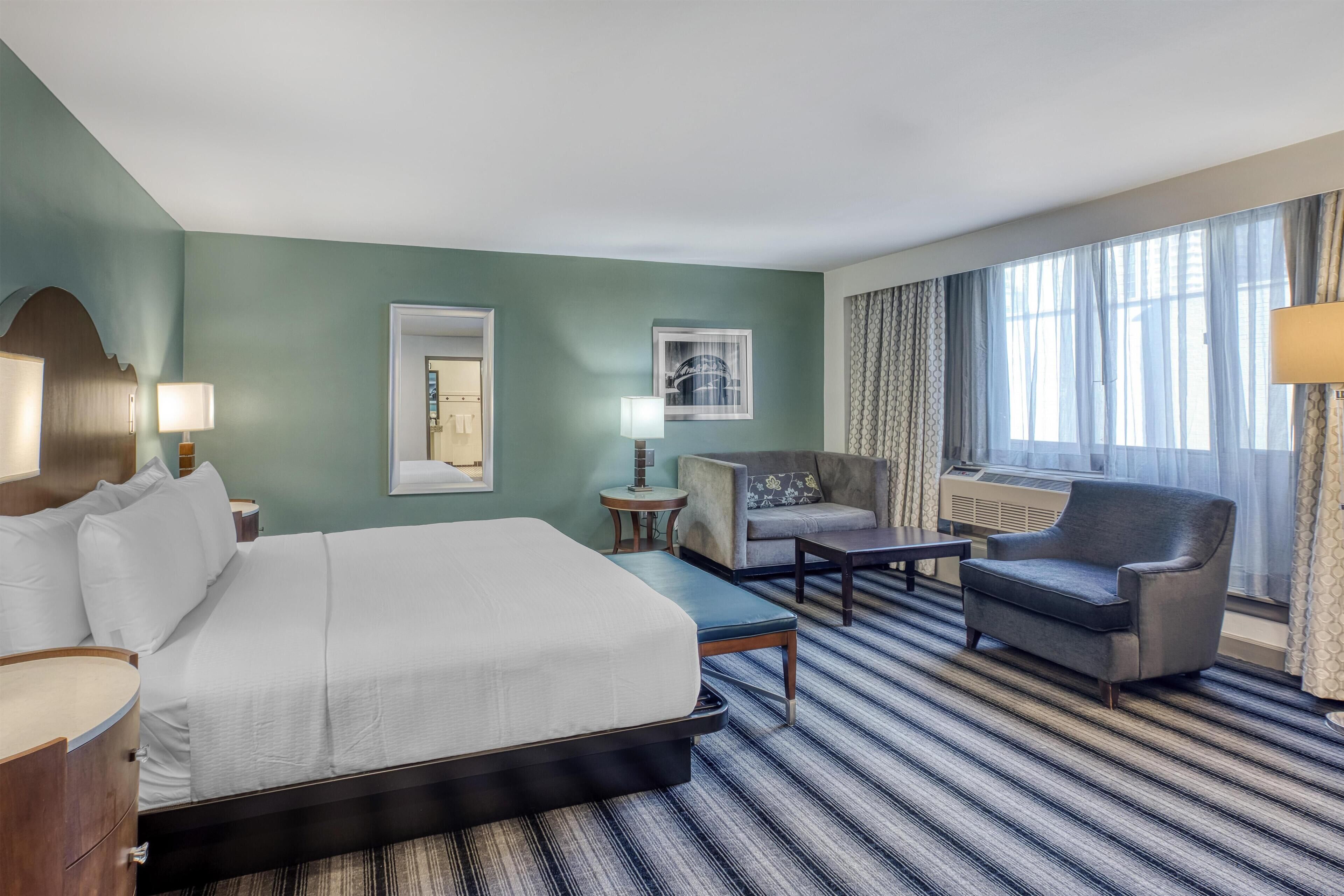 Une chambre d’hôtel avec un grand lit, un canapé, une petite table et une chaise. On y trouve deux lampes, un tableau encadré au mur et une fenêtre avec des rideaux.