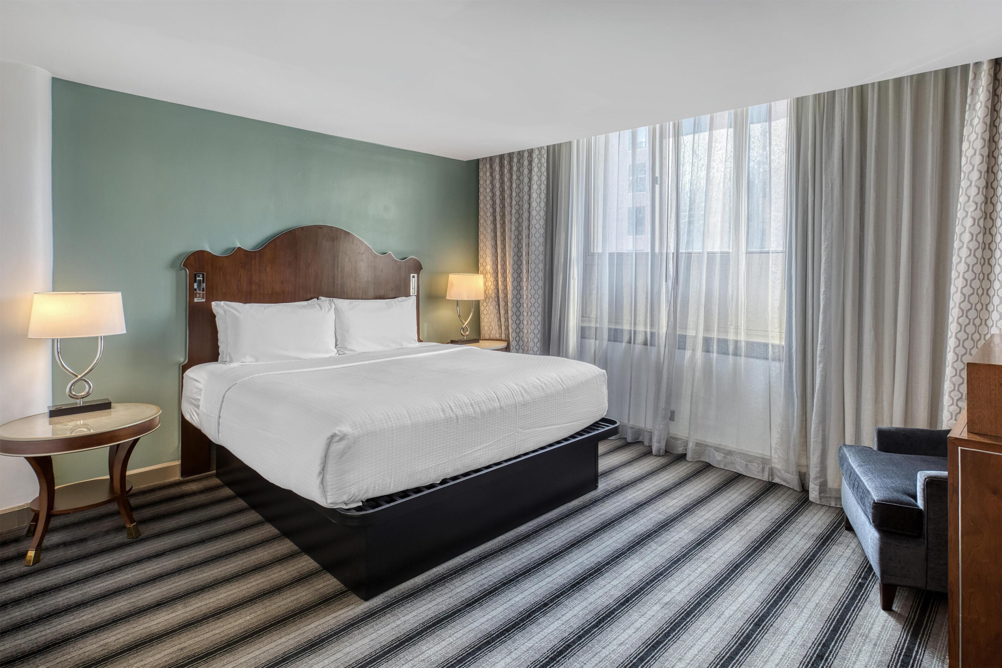 A hotel room with a large bed, a bedside table with a lamp, a small table, a bench, and a large window with sheer curtains.
