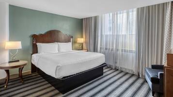 A hotel room with a large bed, a bedside table with a lamp, a small table, a bench, and a large window with sheer curtains.