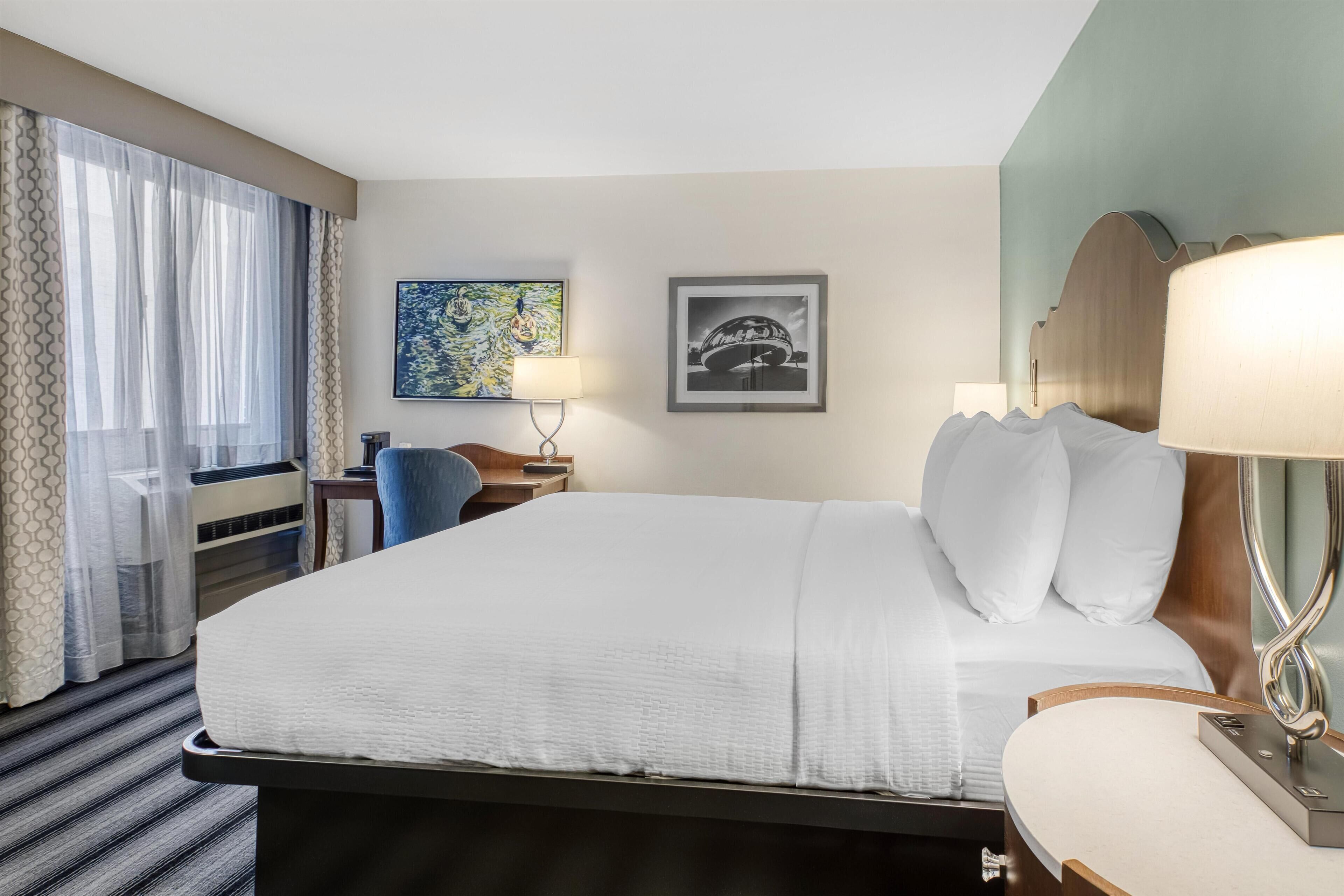 Quarto de hotel com uma cama grande, uma escrivaninha, uma cadeira, um abajur e duas imagens emolduradas na parede.