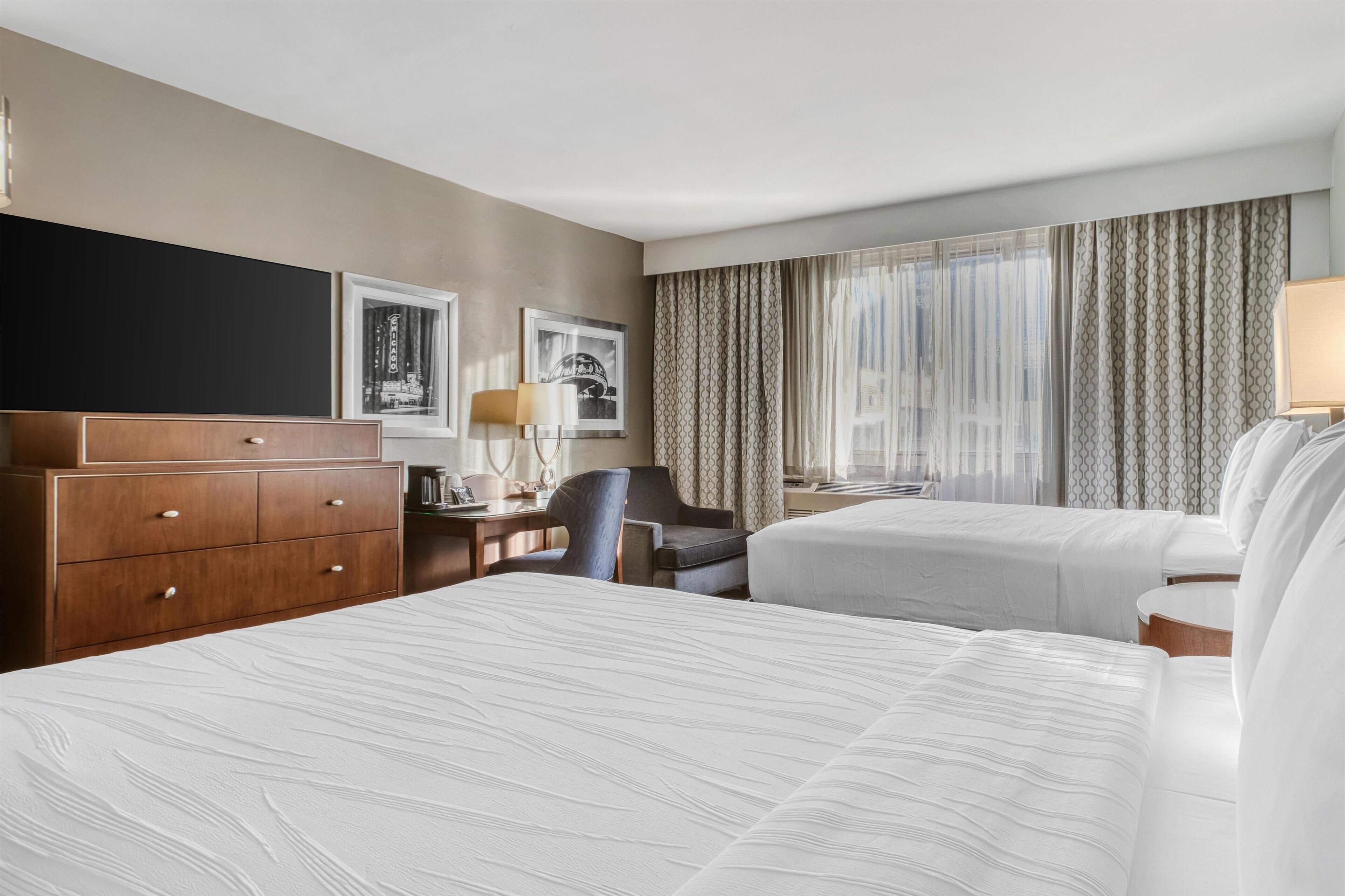 Ein Hotelzimmer mit zwei Betten, einem Schreibtisch, einem Stuhl, einem Schminktisch und einem Fenster mit Vorhängen.