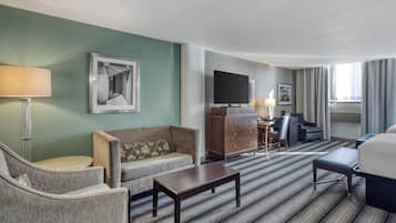 A hotel room with a sofa, armchair, coffee table, TV, desk, chair, and a bed.
