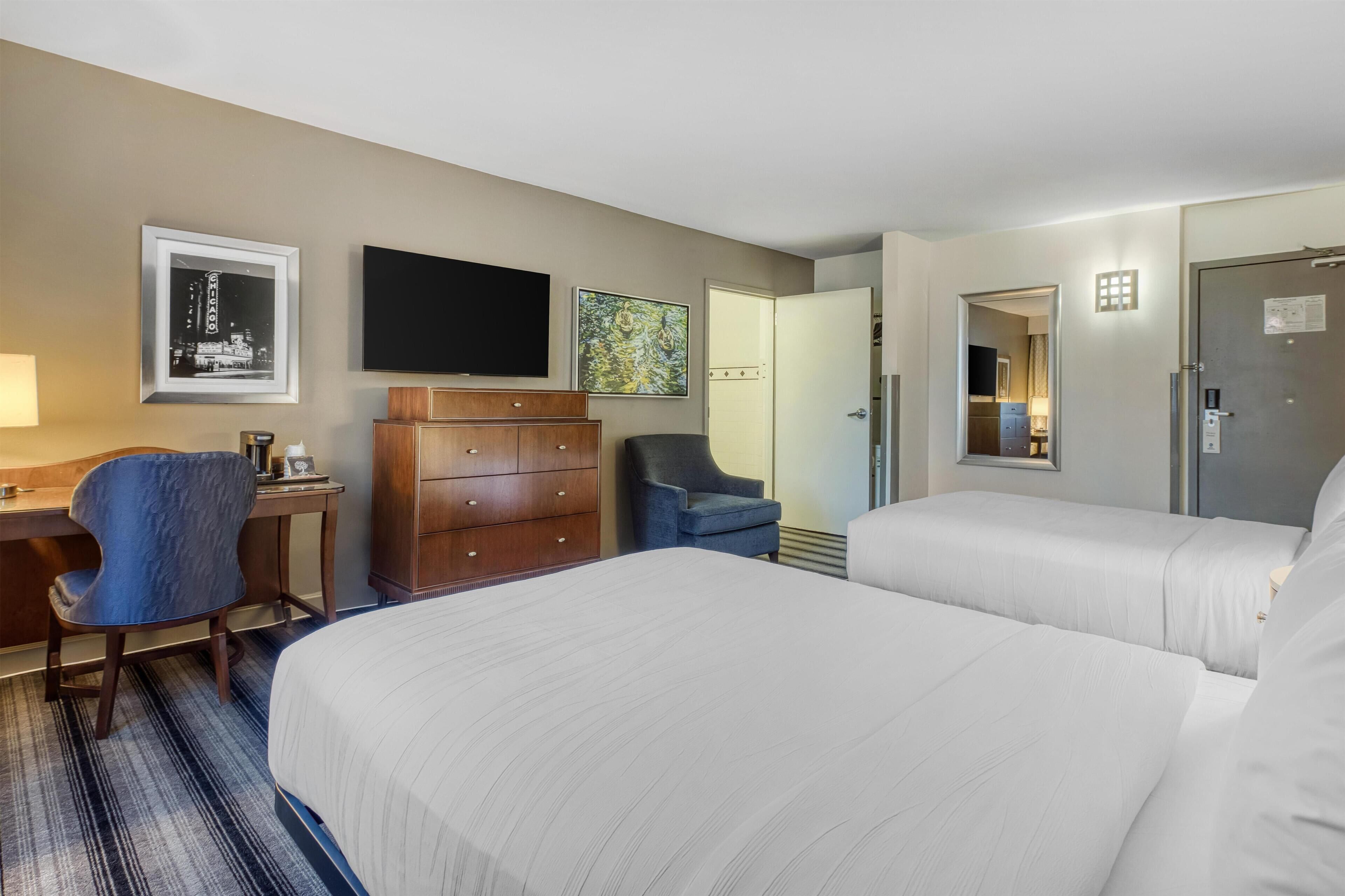 A hotel room with two beds, a desk with a chair, a dresser, a flat-screen TV, and artwork on the walls.