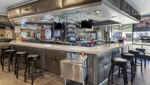 A modern bar area with a stainless steel bar counter, multiple stools, a refrigerated display, and a large window offering a view of the city. - Best Western River North Hotel (Chicago)