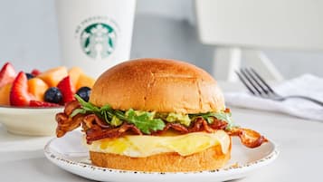 Un sándwich de desayuno con tocino, lechuga y queso en un pan, un plato de fruta y una taza Starbucks al fondo.
