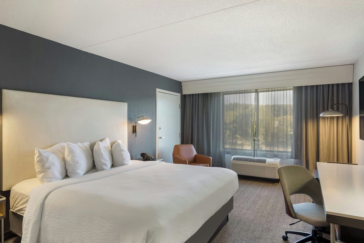 A hotel room with a large bed, a desk, a chair, a phone, a lamp, and a window with curtains.