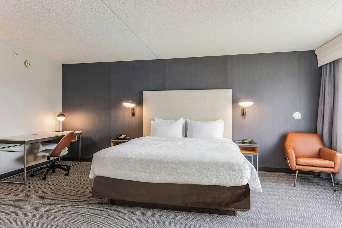 A hotel room with a large bed, a desk with a chair, a nightstand, and a single orange chair.