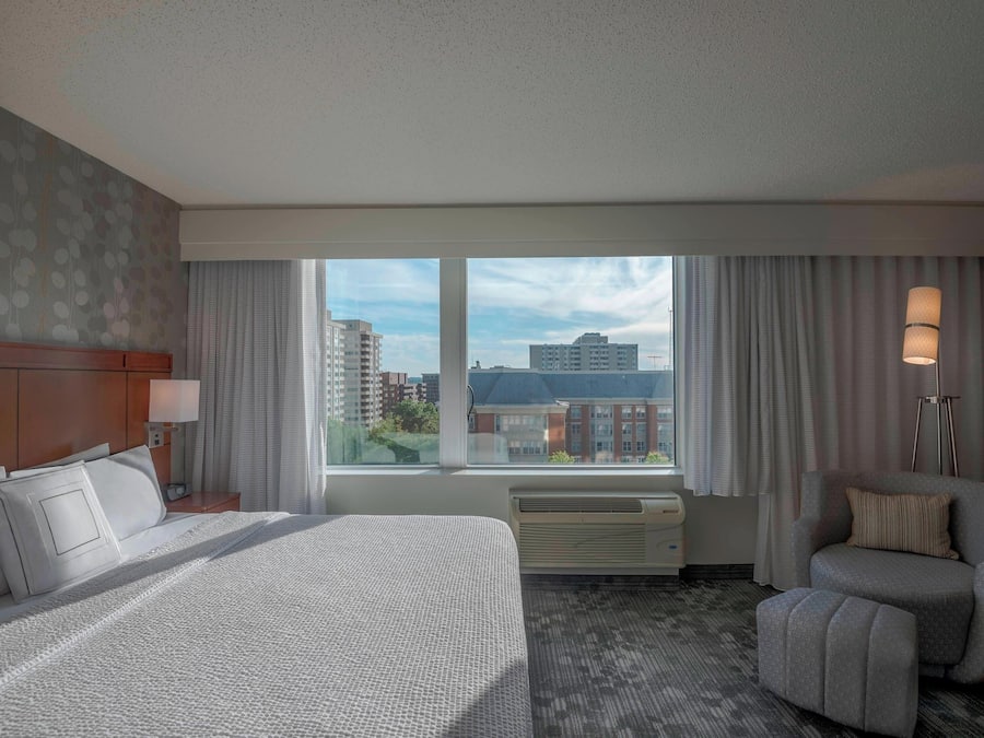 Een hotelkamer met een groot bed, een stoel, een bureau met een lamp en uitzicht op gebouwen door het raam.