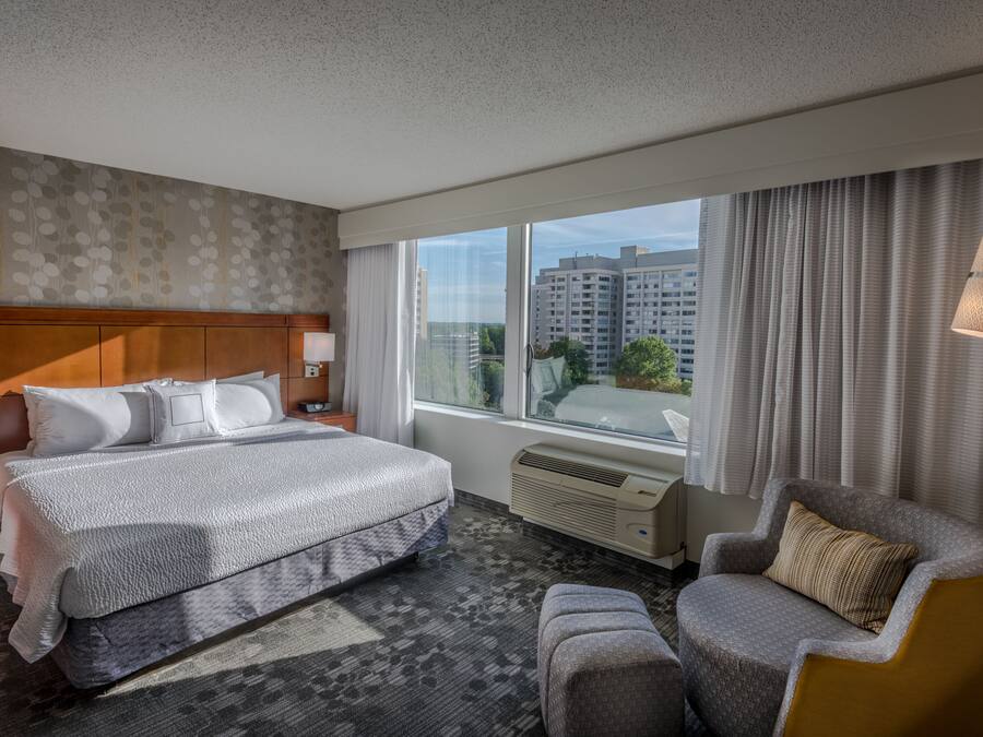 A hotel room with a large bed, a chair, a nightstand, a lamp, a window, and a view of buildings.