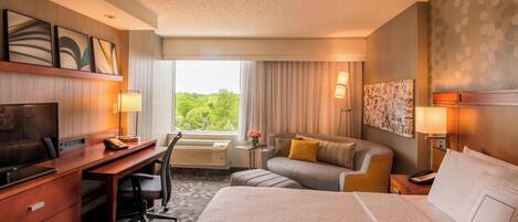Habitación de hotel con cama, escritorio, silla y un ventanal con vistas a zonas verdes.