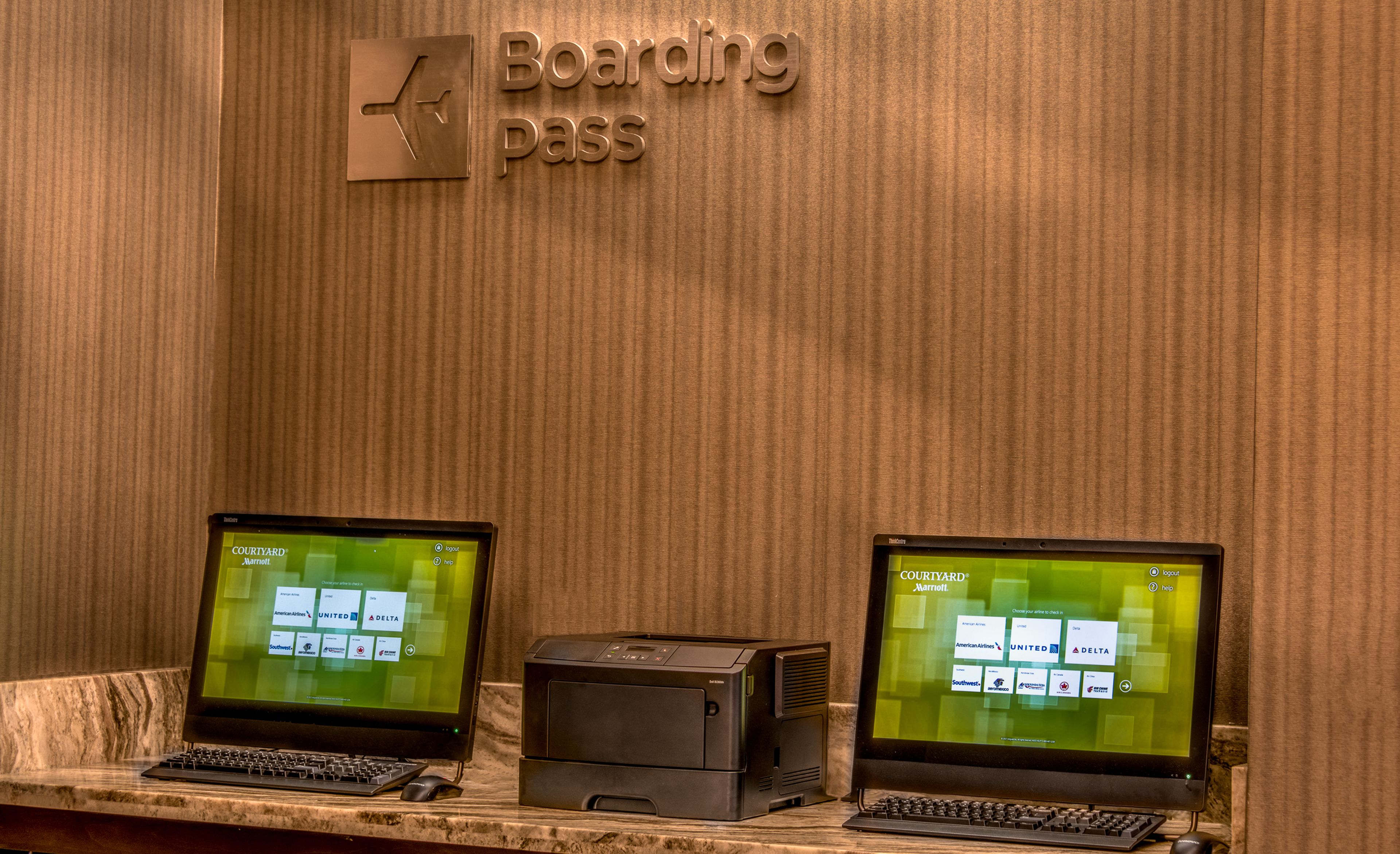 Two computer monitors displaying airline check-in screens, a printer, and a keyboard on a counter with a 'Boarding Pass' sign above.