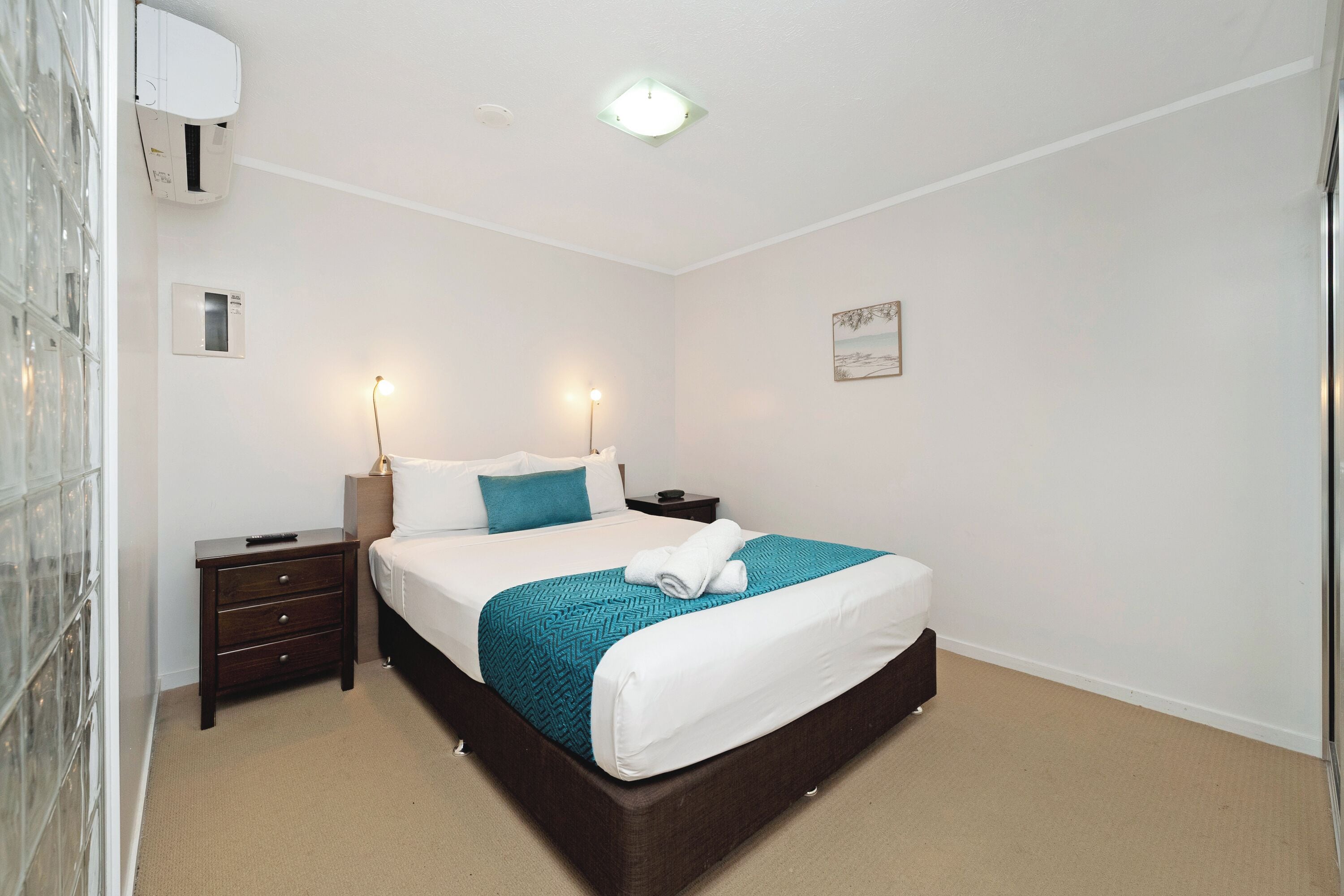A neatly arranged bedroom with a bed, bedside tables, a nightstand, a wall-mounted air conditioner, and a ceiling light.