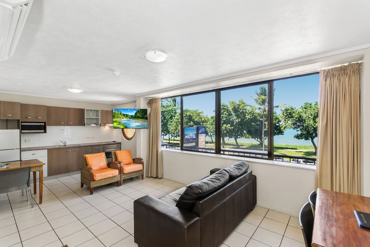 A modern hotel room with a kitchen, a sofa, a dining table, and a large window with a view of a tropical landscape.
