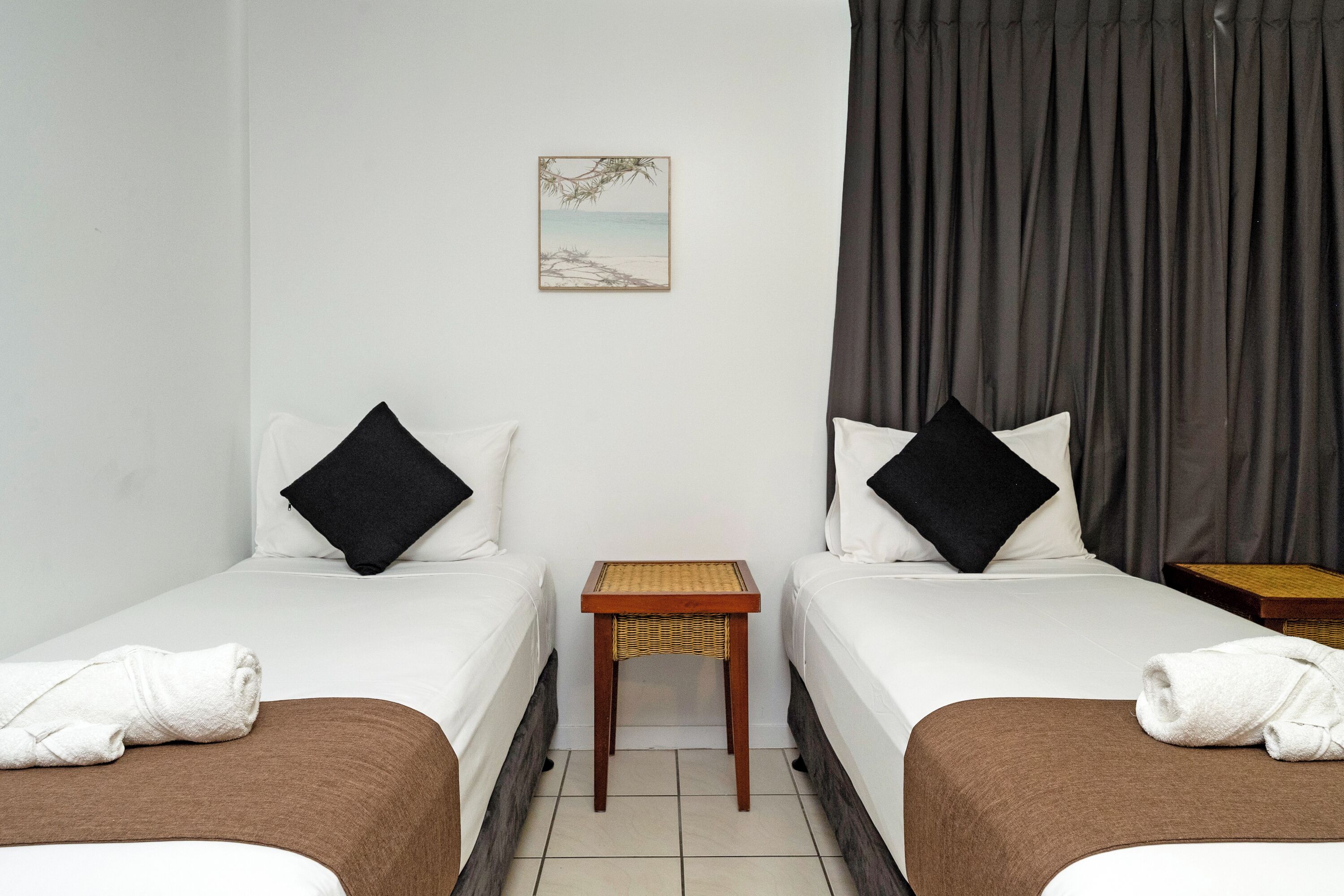 A hotel room with two beds, a small table, a picture on the wall, and a curtain.