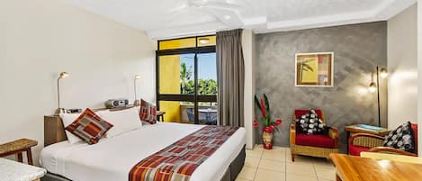 Habitación de hotel con una cama grande, un escritorio, una silla, una mesita, un ventilador de techo, una ventana con cortinas y una lámpara de pared.