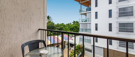 A balcony with a glass table and chairs, overlooking a swimming pool and adjacent buildings.