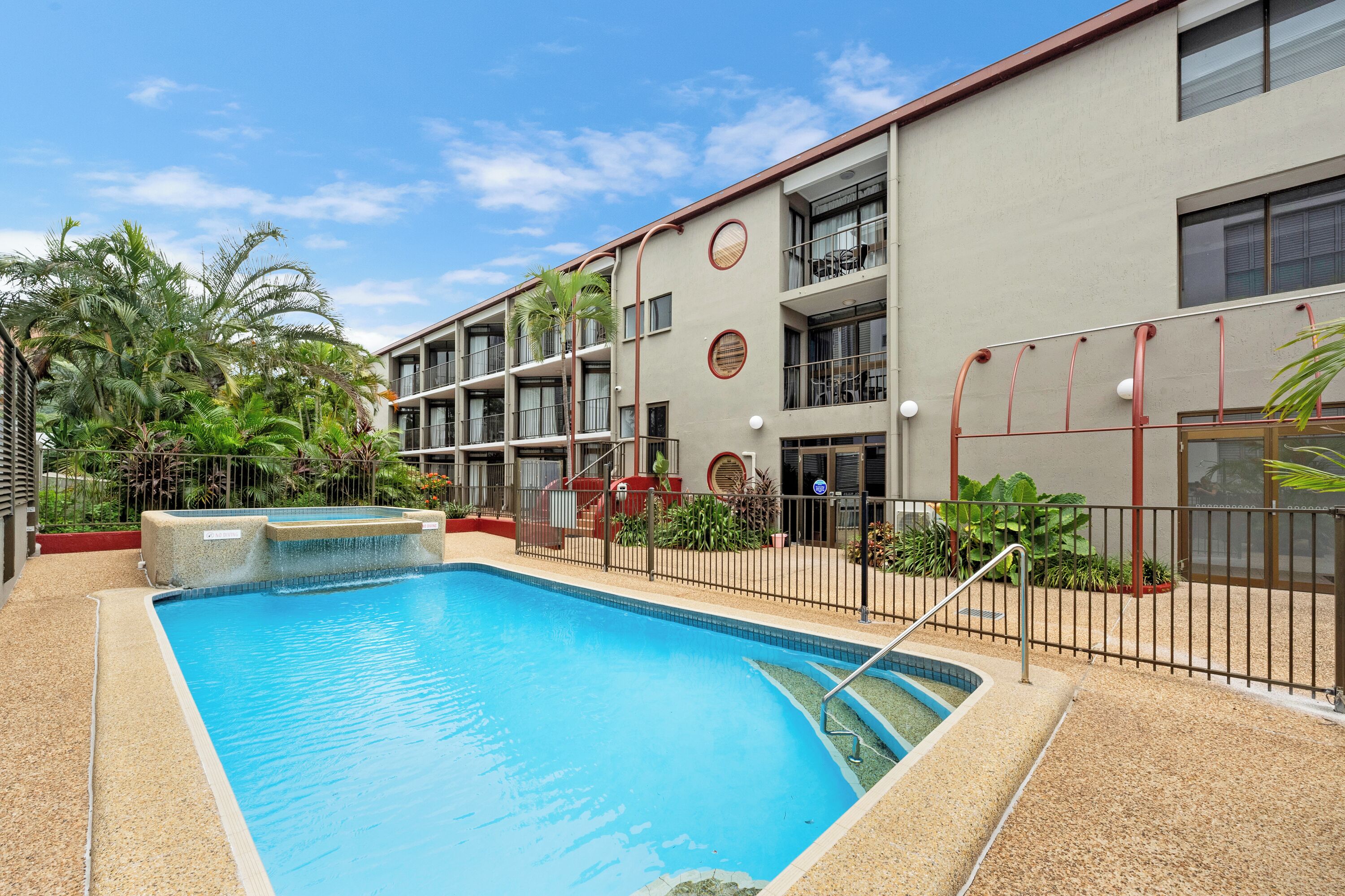 A swimming pool with a surrounding fence, a building with multiple balconies, and tropical plants.