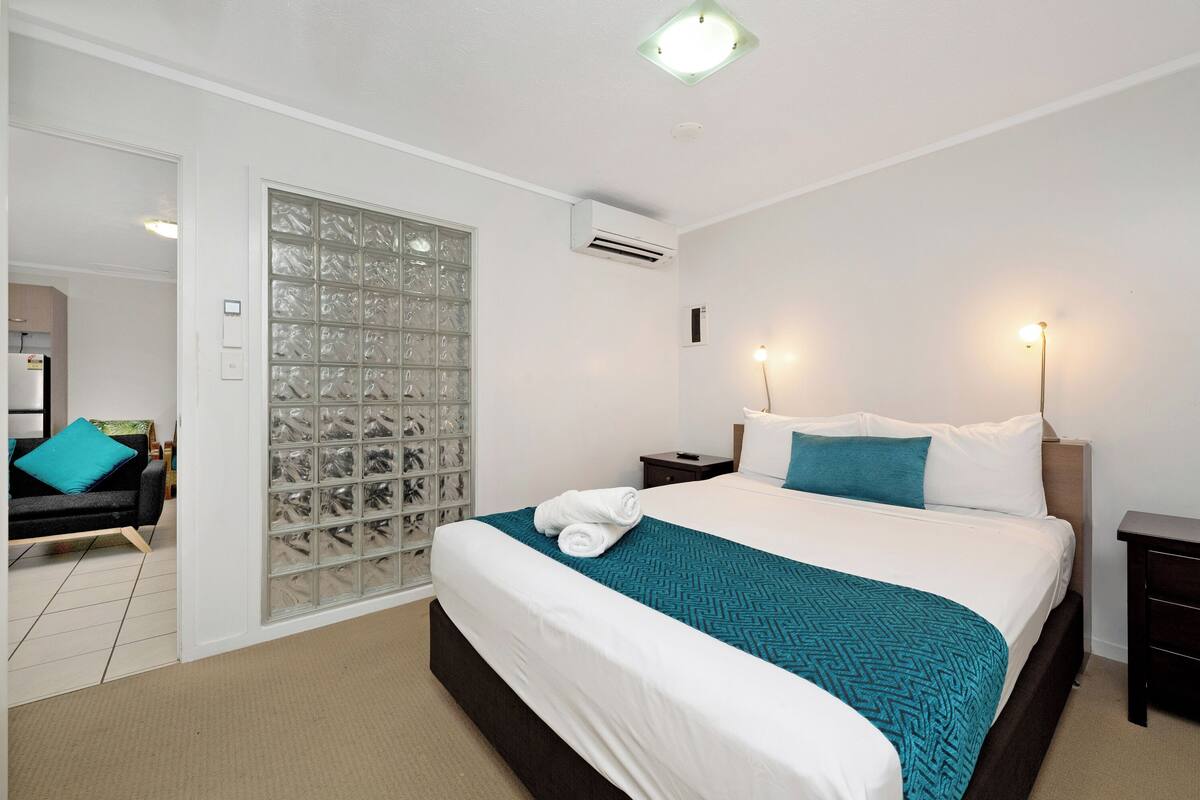 A hotel room with a bed, a nightstand, a wall-mounted air conditioner, and a sliding glass door leading to a bathroom.