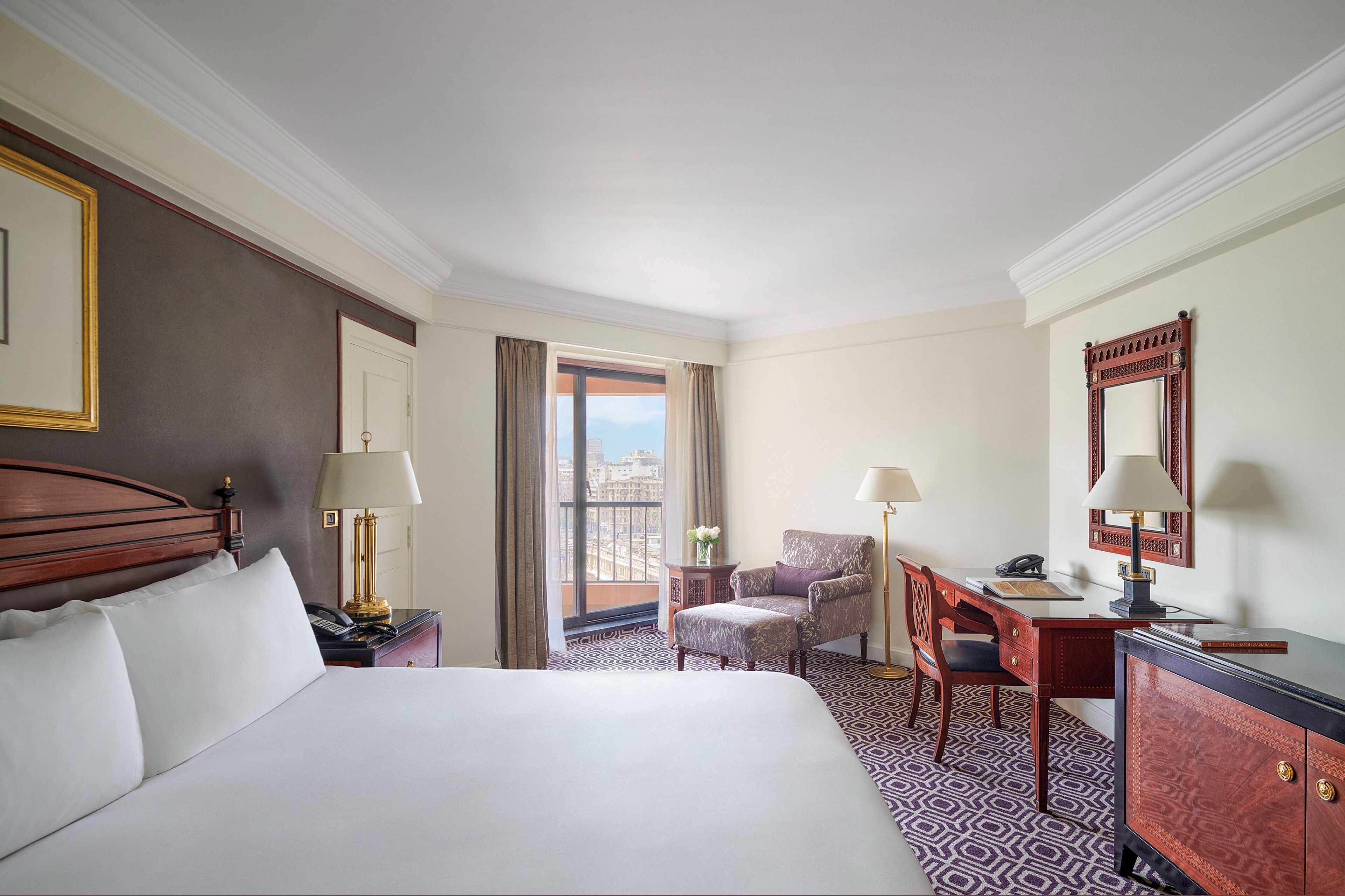 A hotel room with a large bed, a desk with a chair, a small table with a lamp, and a large mirror.