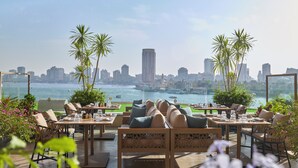 Un espace extérieur aménagé avec des tables et des chaises, offrant une vue sur un plan d’eau et le panorama de la ville.