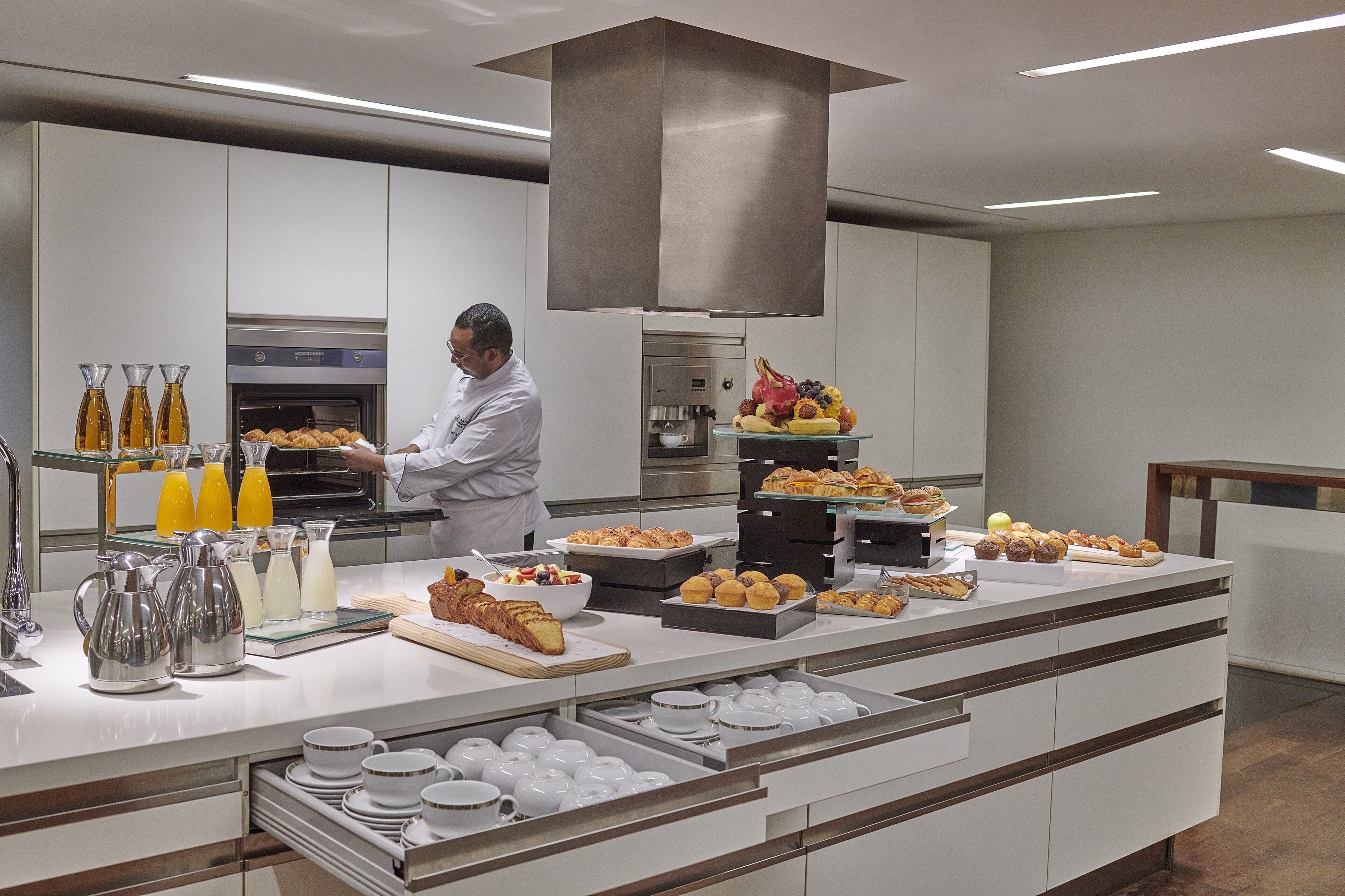 Een moderne keuken met een kok die eten bereidt, een buffet met diverse gebakjes en fruit, en een koffiehoek met meerdere koffiezetapparaten en kopjes.