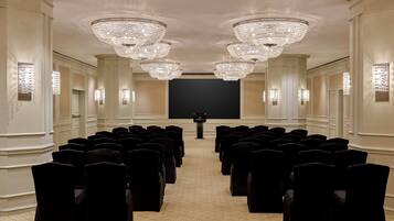 A conference room with rows of chairs facing a large screen, ornate chandeliers, and wall sconces.