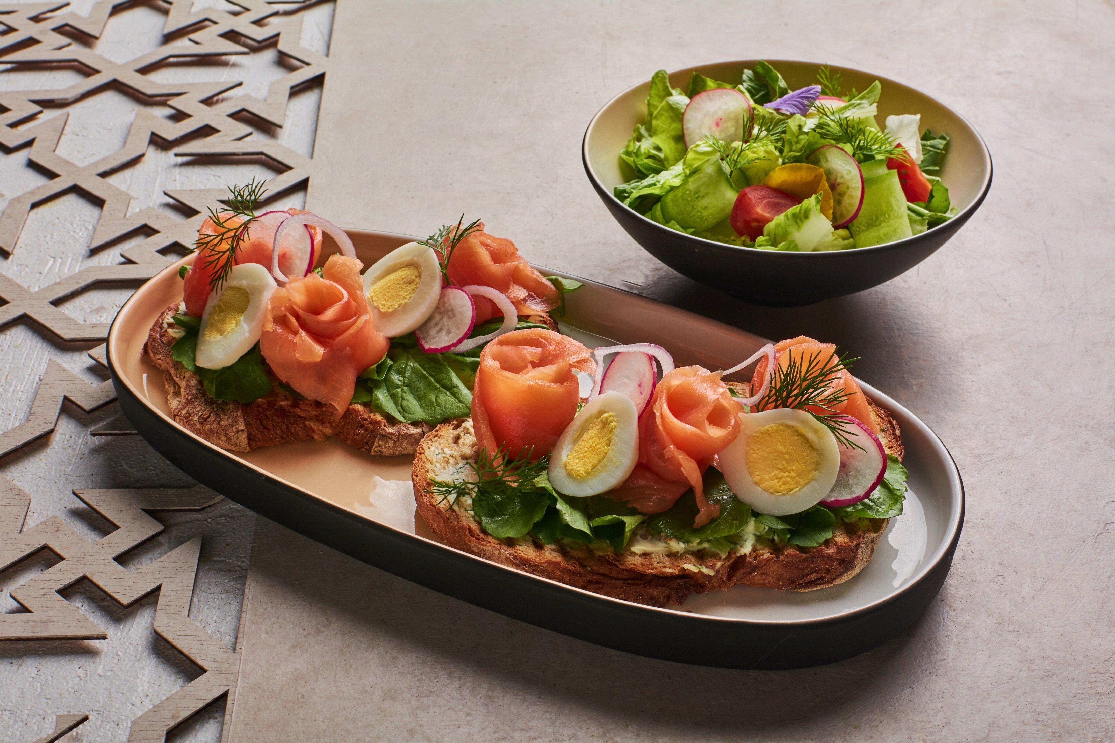A plate with two open-faced sandwiches topped with smoked salmon, eggs, and greens, and a bowl of mixed greens salad.