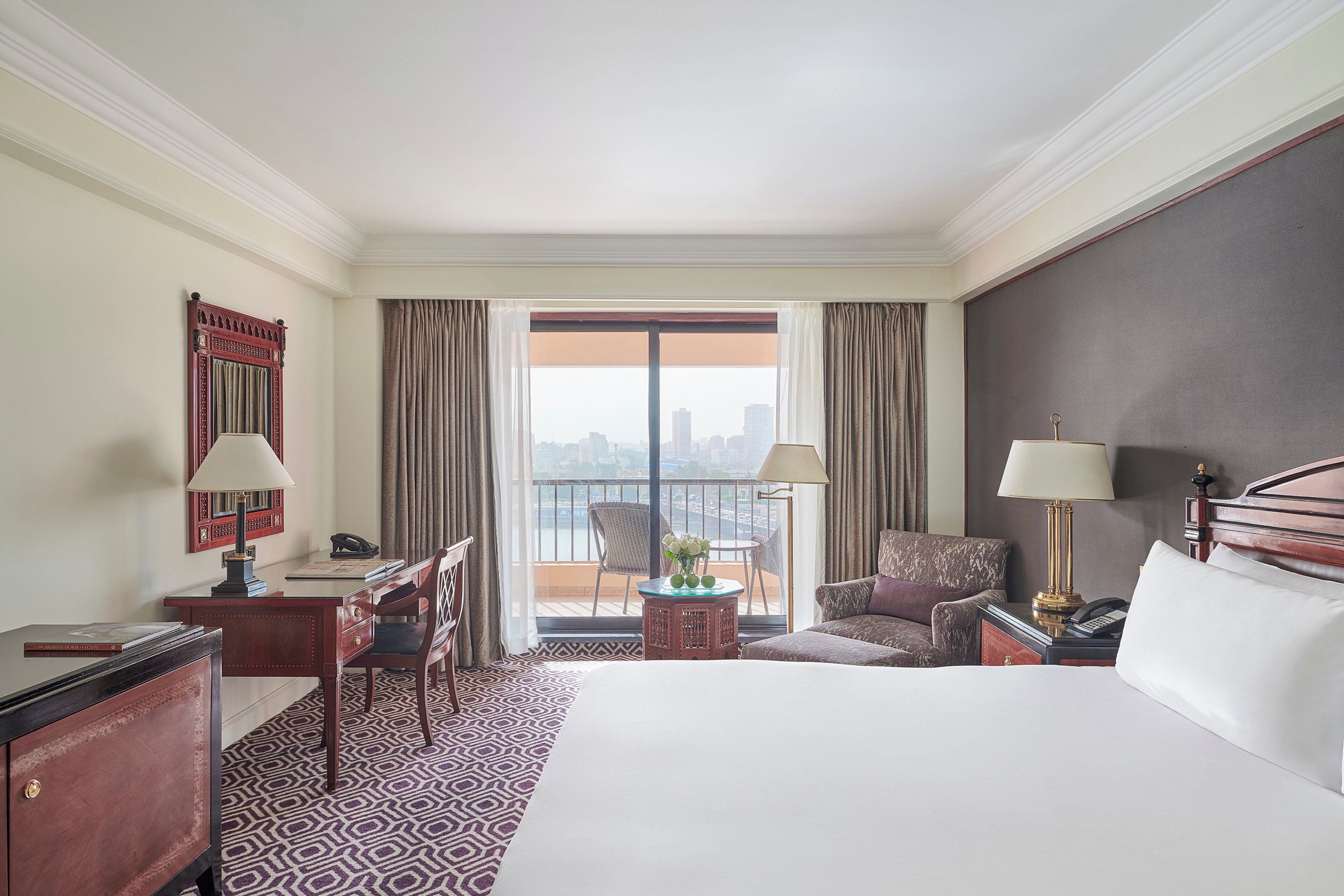 A hotel room with a large bed, a desk with a lamp, a chair, a small table with flowers, and a view of the city through the window.