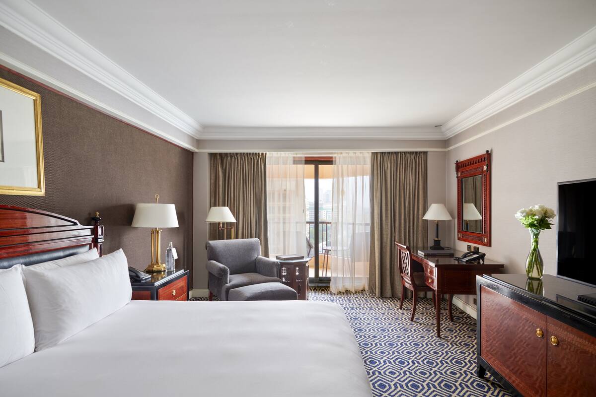 A hotel room with a large bed, a desk, a chair, a TV, and a view of the city through the window.
