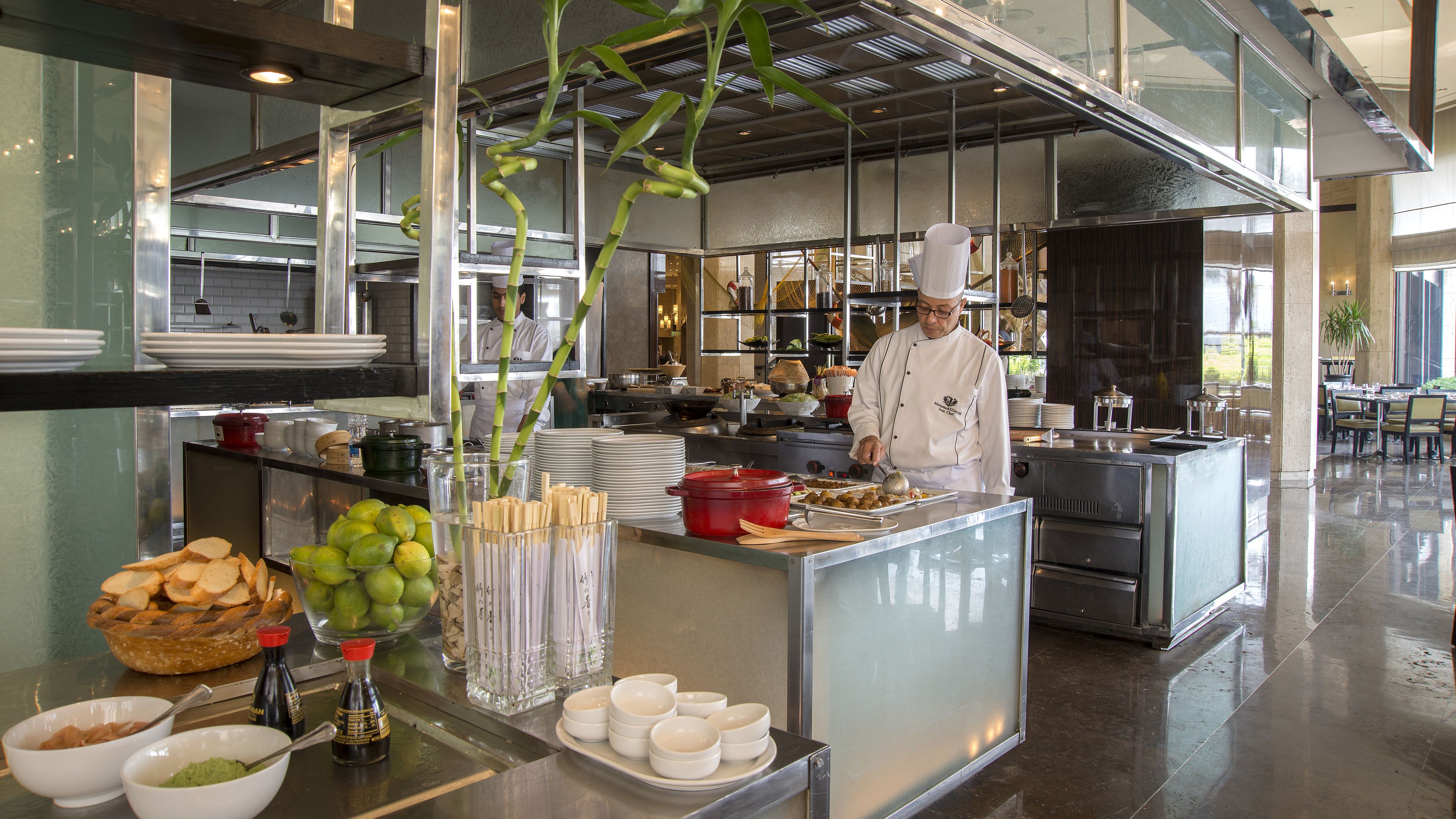 A modern kitchen with chefs preparing food, a buffet area with various dishes and condiments, and a dining area with tables and chairs.