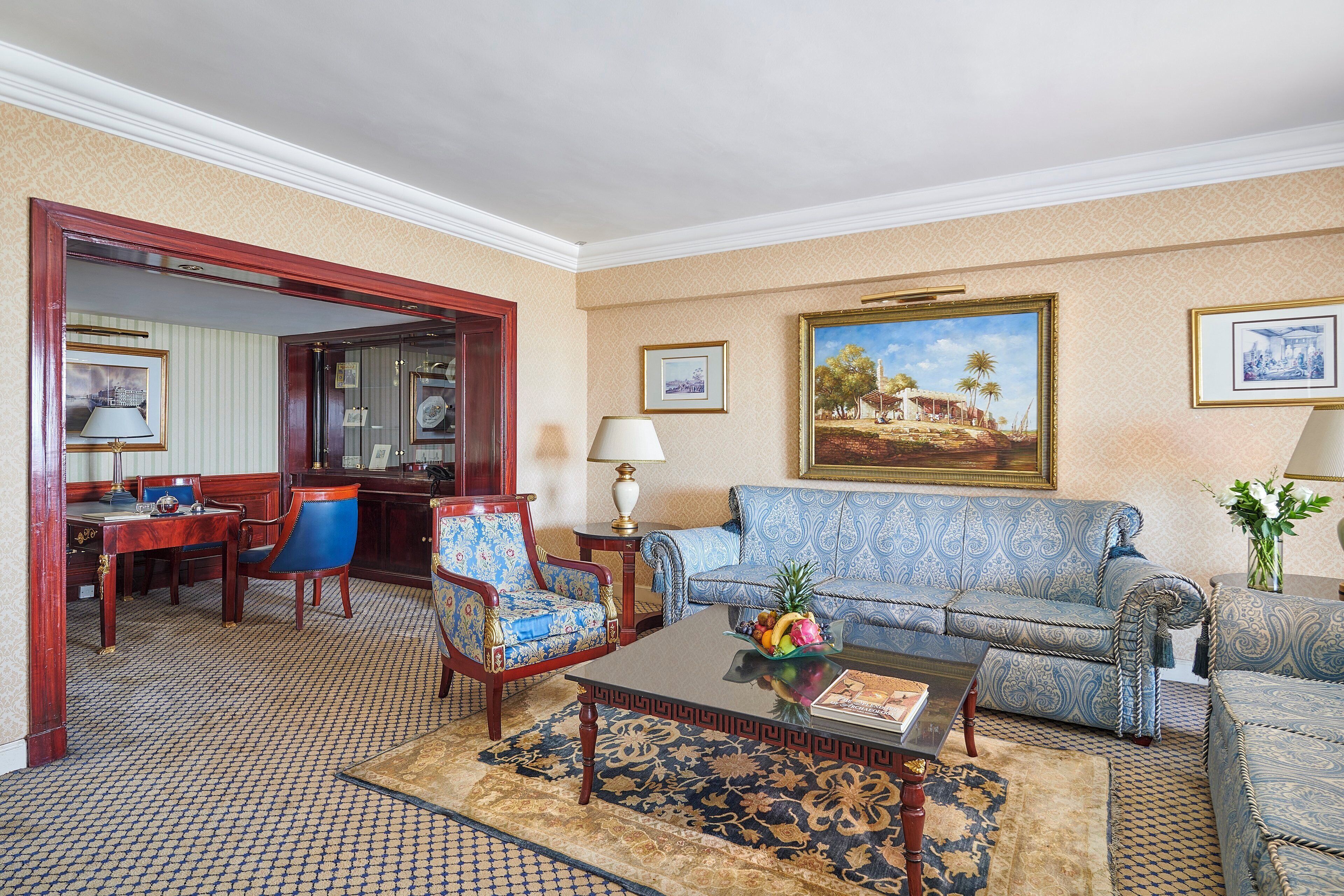 A room with a patterned carpet, a blue sofa, a wooden chair, a coffee table with a fruit bowl, and a painting on the wall.