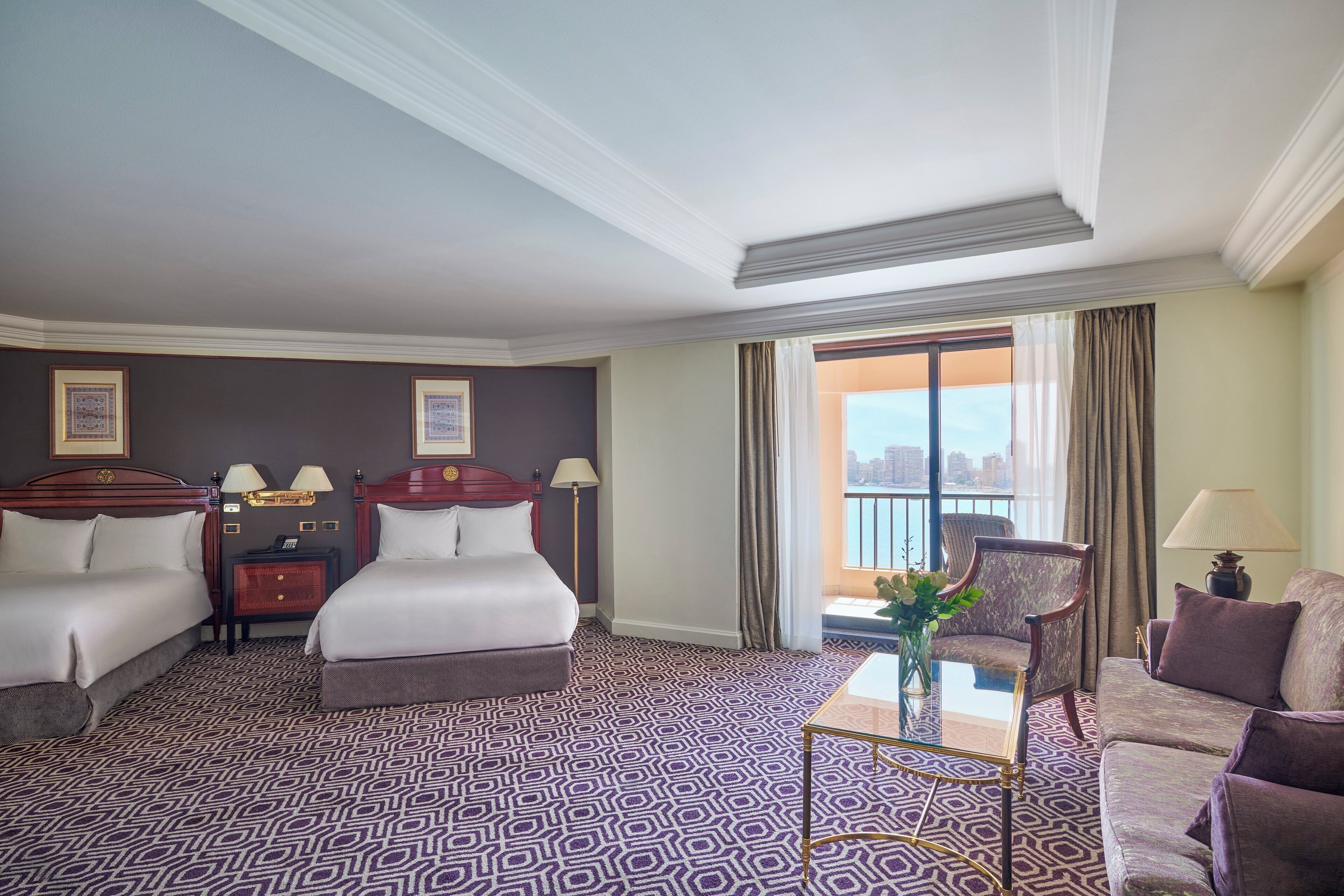 A hotel room with a large bed, two bedside tables with lamps, a seating area with a sofa and armchairs, a patterned carpet, and a view of the city through a large window.