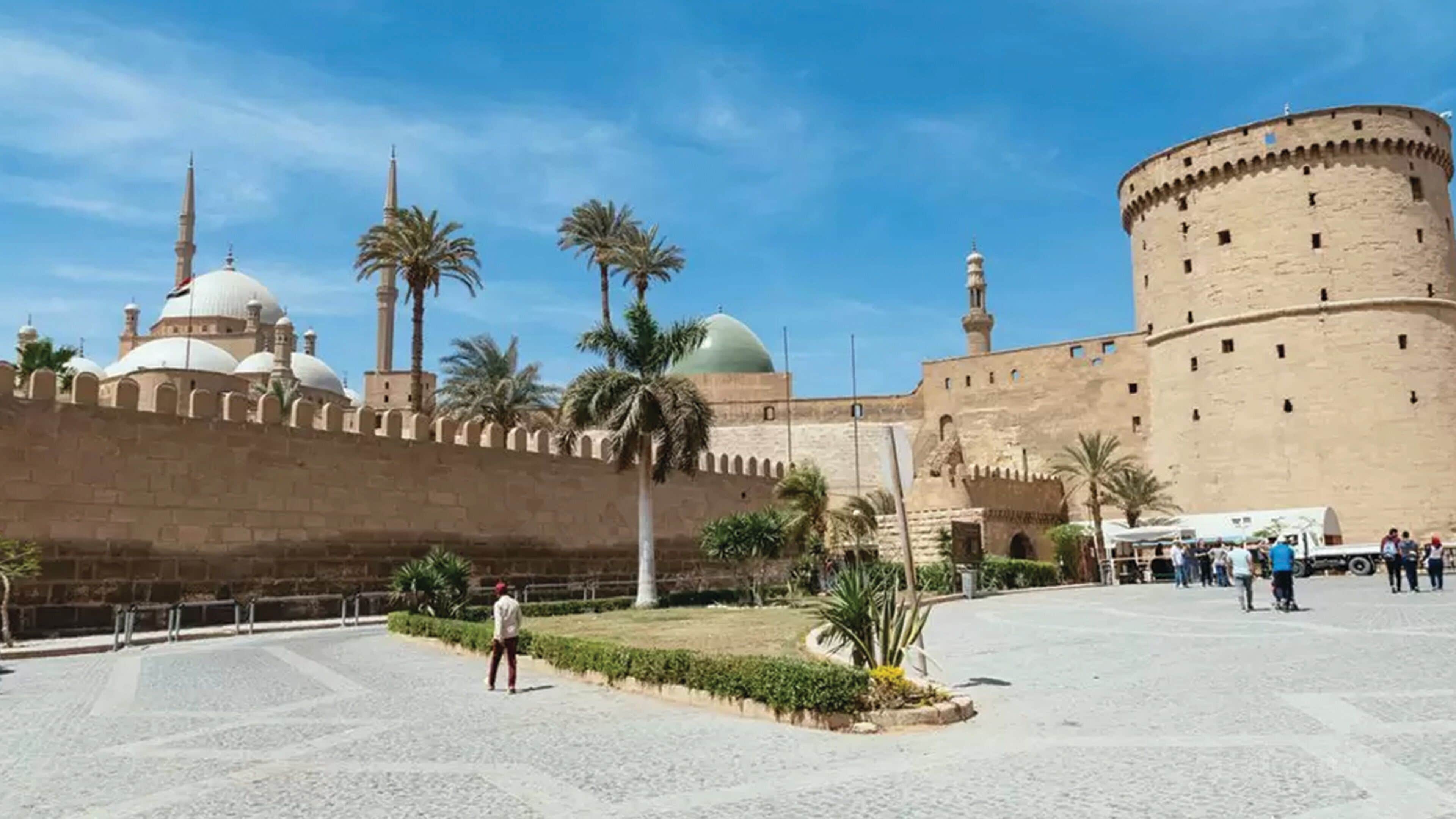 A historical fort with a large central tower, a mosque with domes and minarets, and a paved square with palm trees.