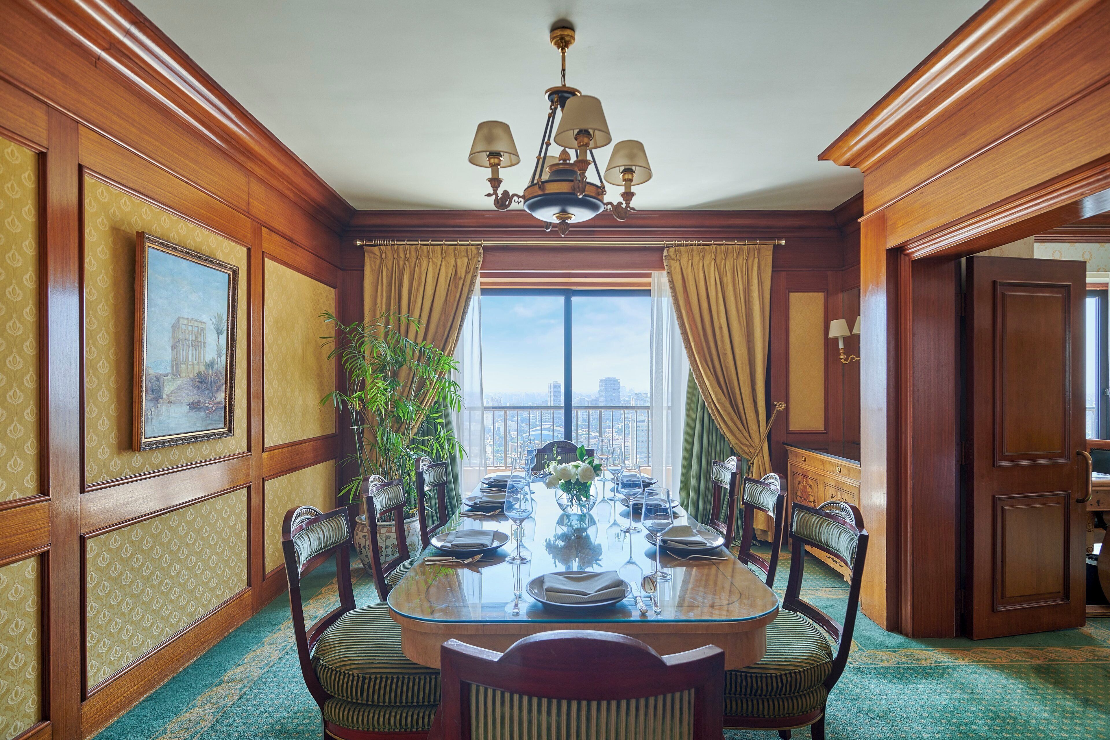 Une salle à manger élégante, avec une grande table dressée pour six personnes, entourée de chaises à l’accoudoir recouvertes d’un tissu à rayures, une grande fenêtre avec des rideaux et une vue sur le paysage urbain.