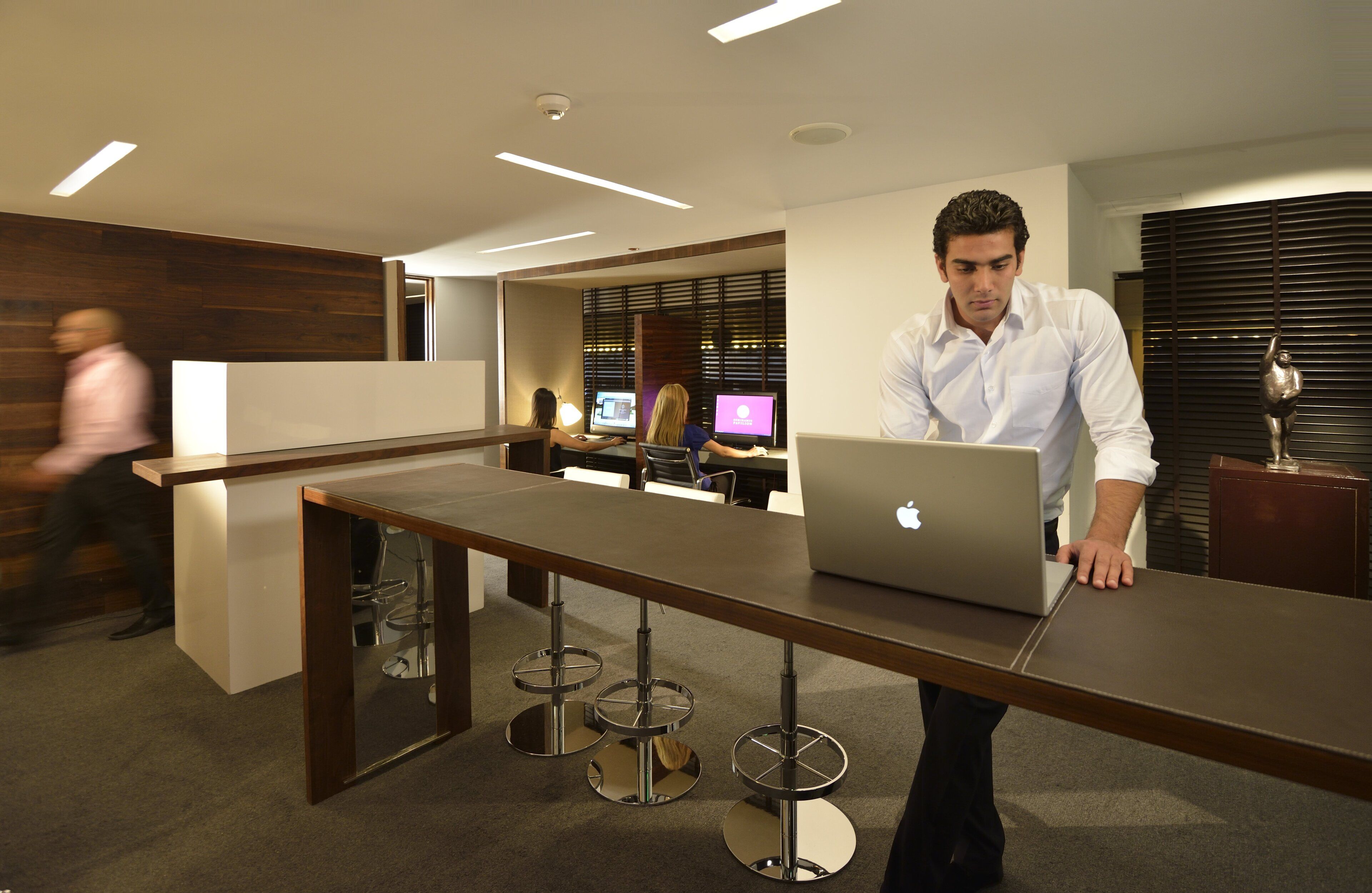 A modern office with a long table, bar stools, and people working at computers.