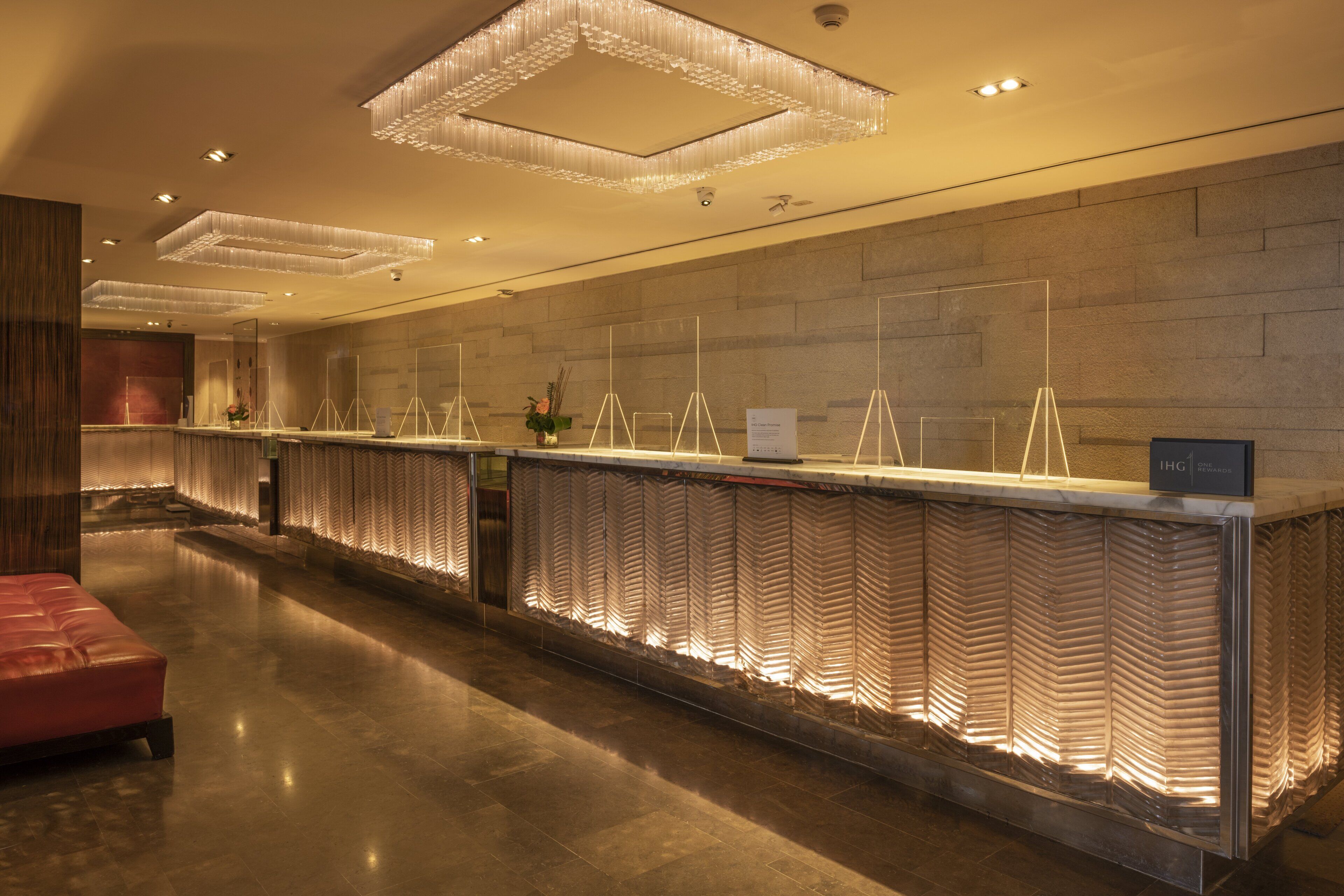 Un'accogliente area reception di un hotel moderno, con un lungo bancone, illuminazione decorativa e una zona salotto in velluto rosso.