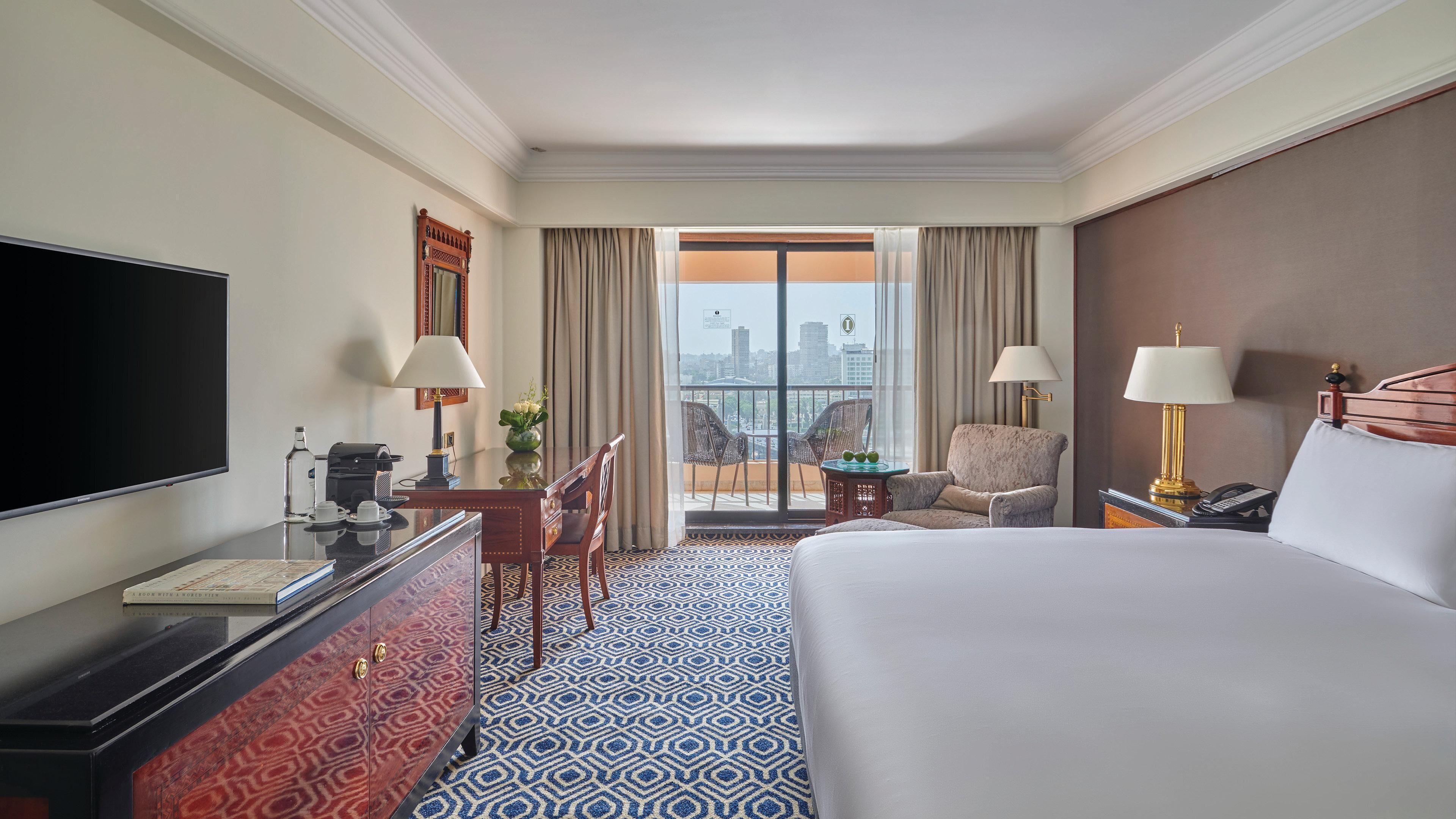 A hotel room with a large bed, a desk with a flat-screen TV, a coffee machine, and a chair. There is a balcony with a view of the city.
