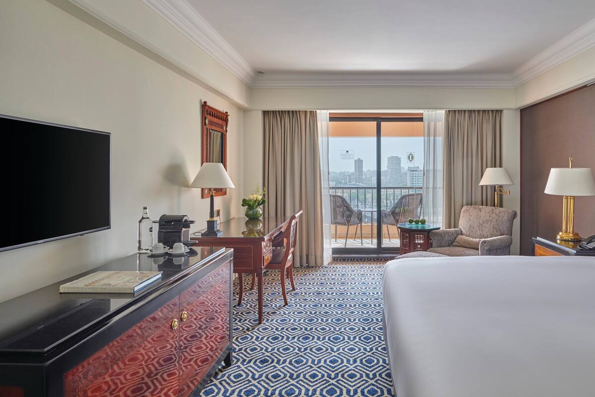 A hotel room with a large bed, a desk with a flat-screen TV, a coffee machine, and a chair. There is a balcony with a view of the city.