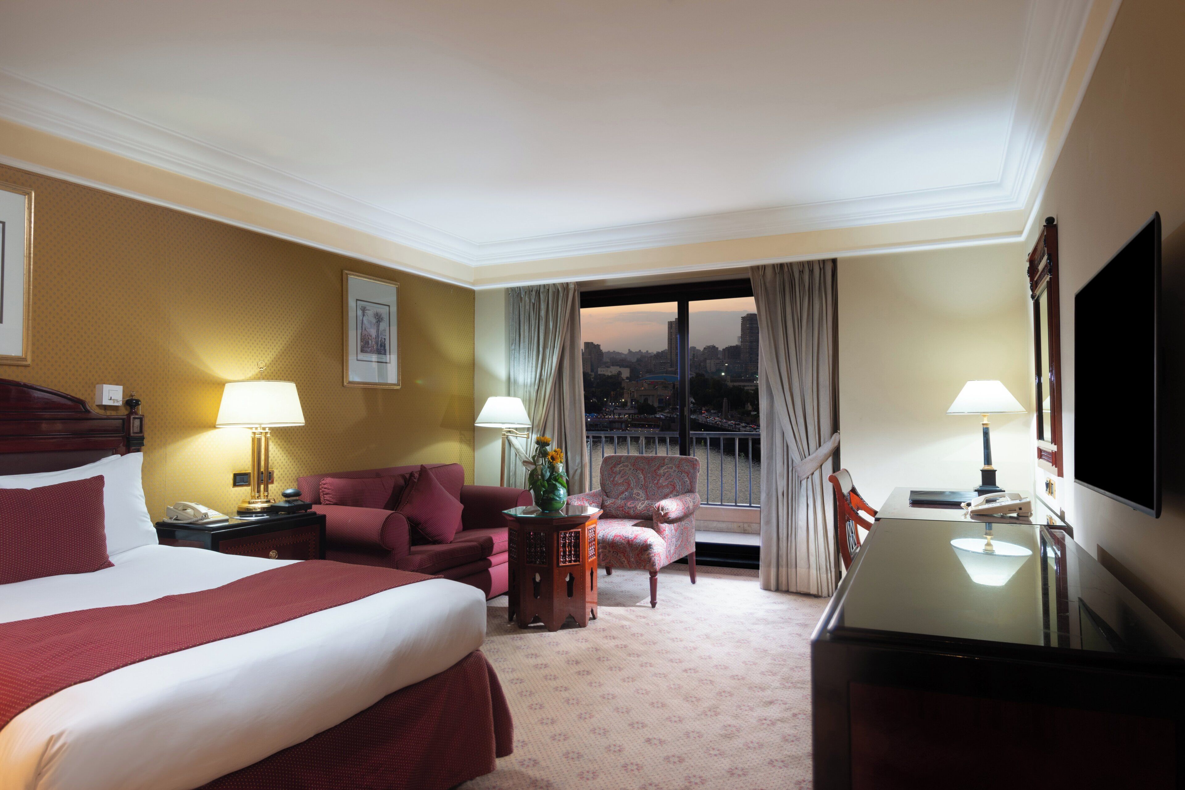 A hotel room with a large bed, a desk, a chair, and a view of the cityscape through the window.