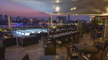 An outdoor dining area with tables and chairs, overlooking a river with city lights in the background.
