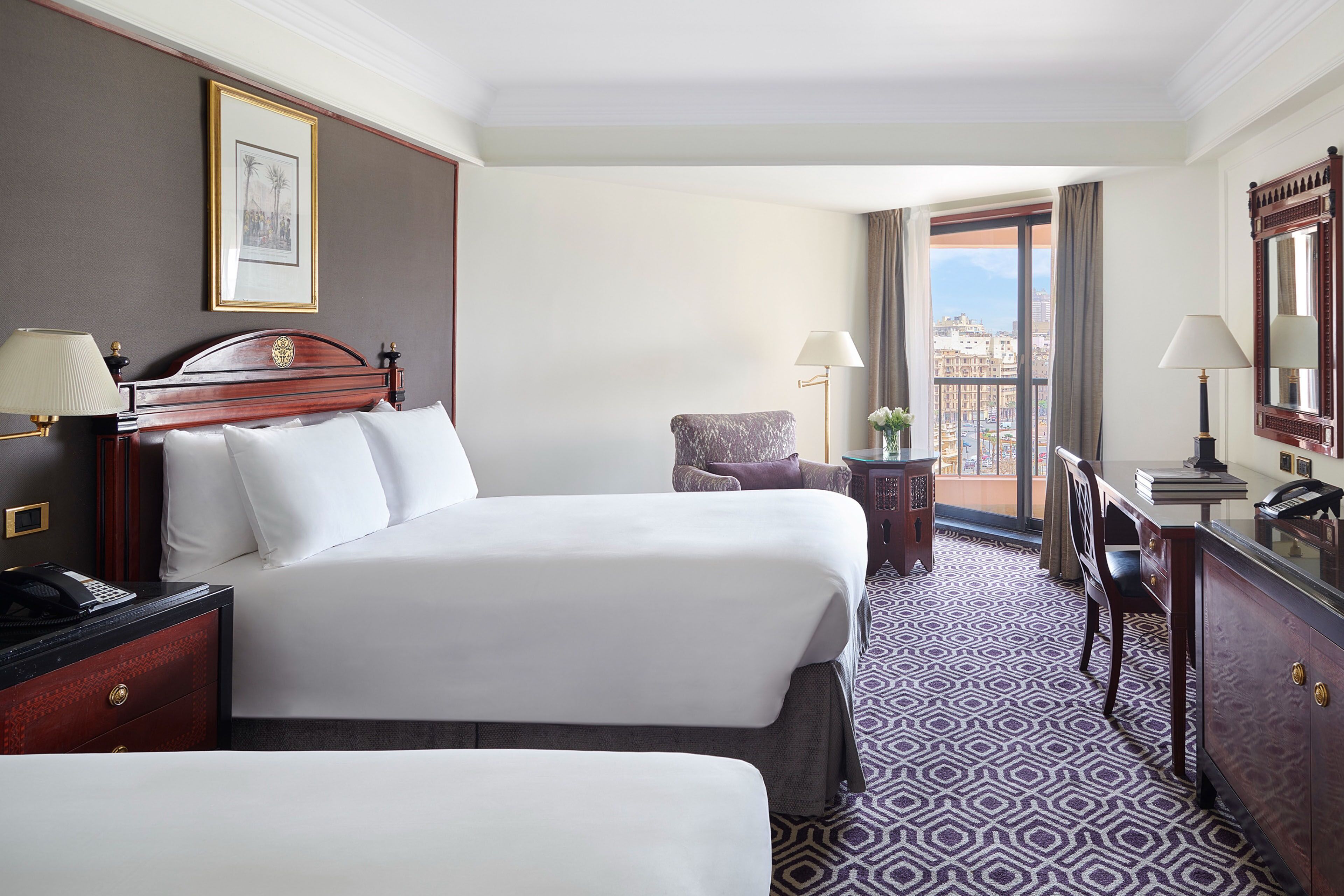 A hotel room with two beds, a desk, a chair, and a balcony with a city view.