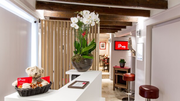 A reception area with a white reception desk, a wicker basket with a teddy bear and a red sign, a potted plant with white orchids, and a red sign with "Dwin" on it. - D'win Hotel (Paris)