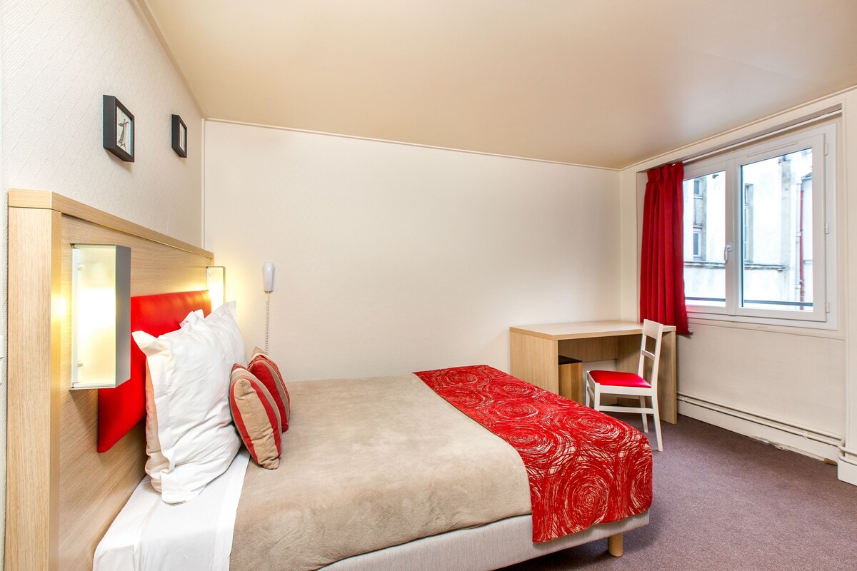 A bedroom with a bed, bedside table, chair, and a window with curtains.